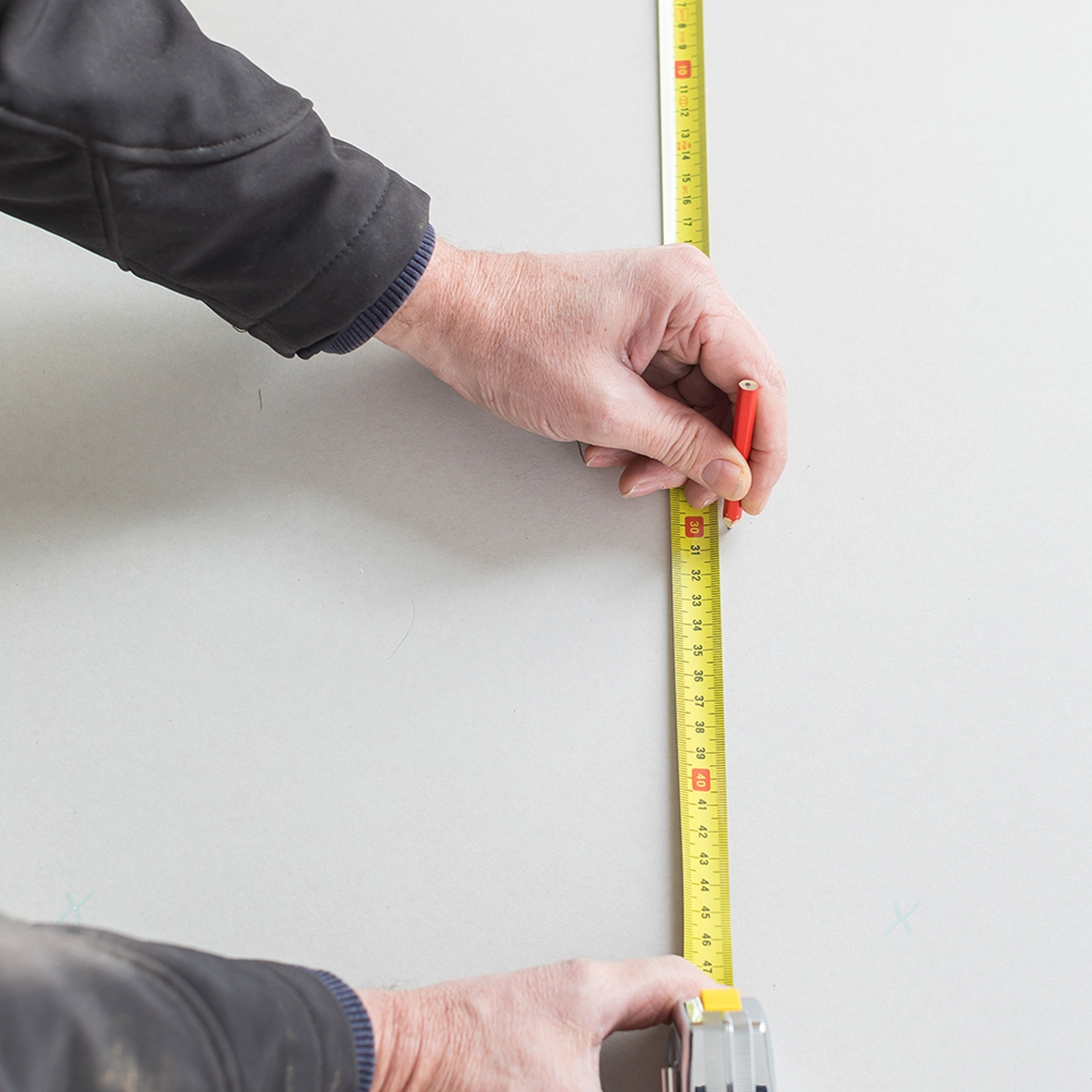 Osoba używa miarki i ołówka do zaznaczania na ścianie. Ściana jest biała.
