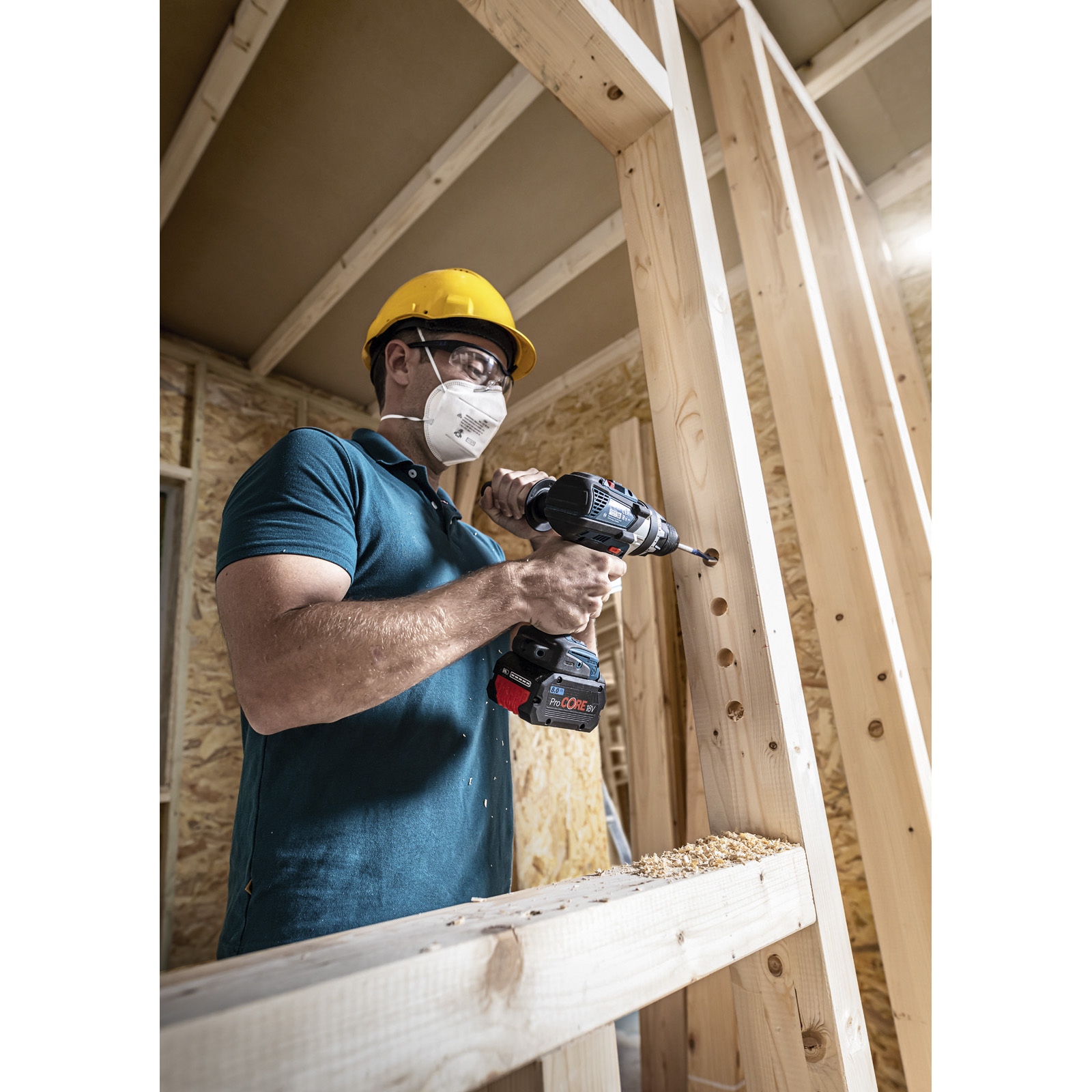 Mężczyzna w kasku i masce wierci w drewnianych słupkach. Scena ma miejsce w pomieszczeniu, podczas budowy.