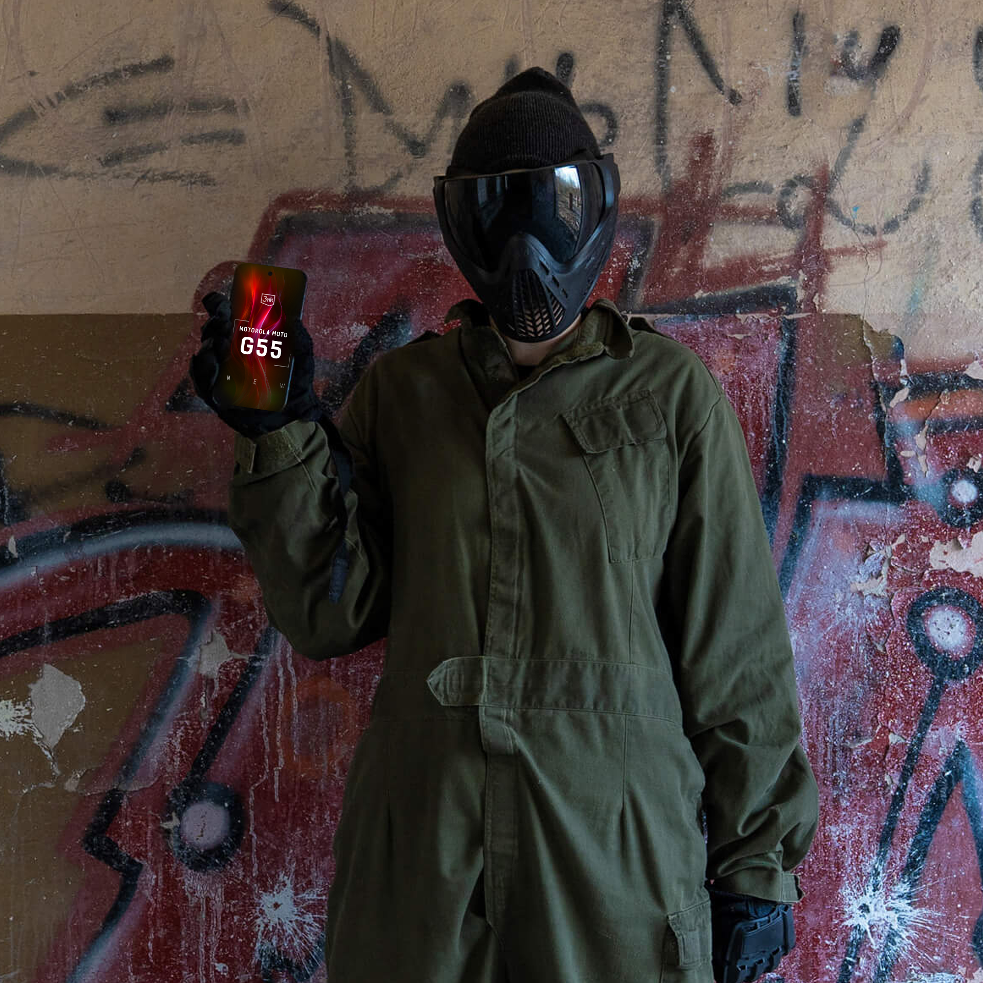 Persona in tuta verde e maschera che tiene un telefono con la scritta "Motorola Moto G55" contro un muro con graffiti.
