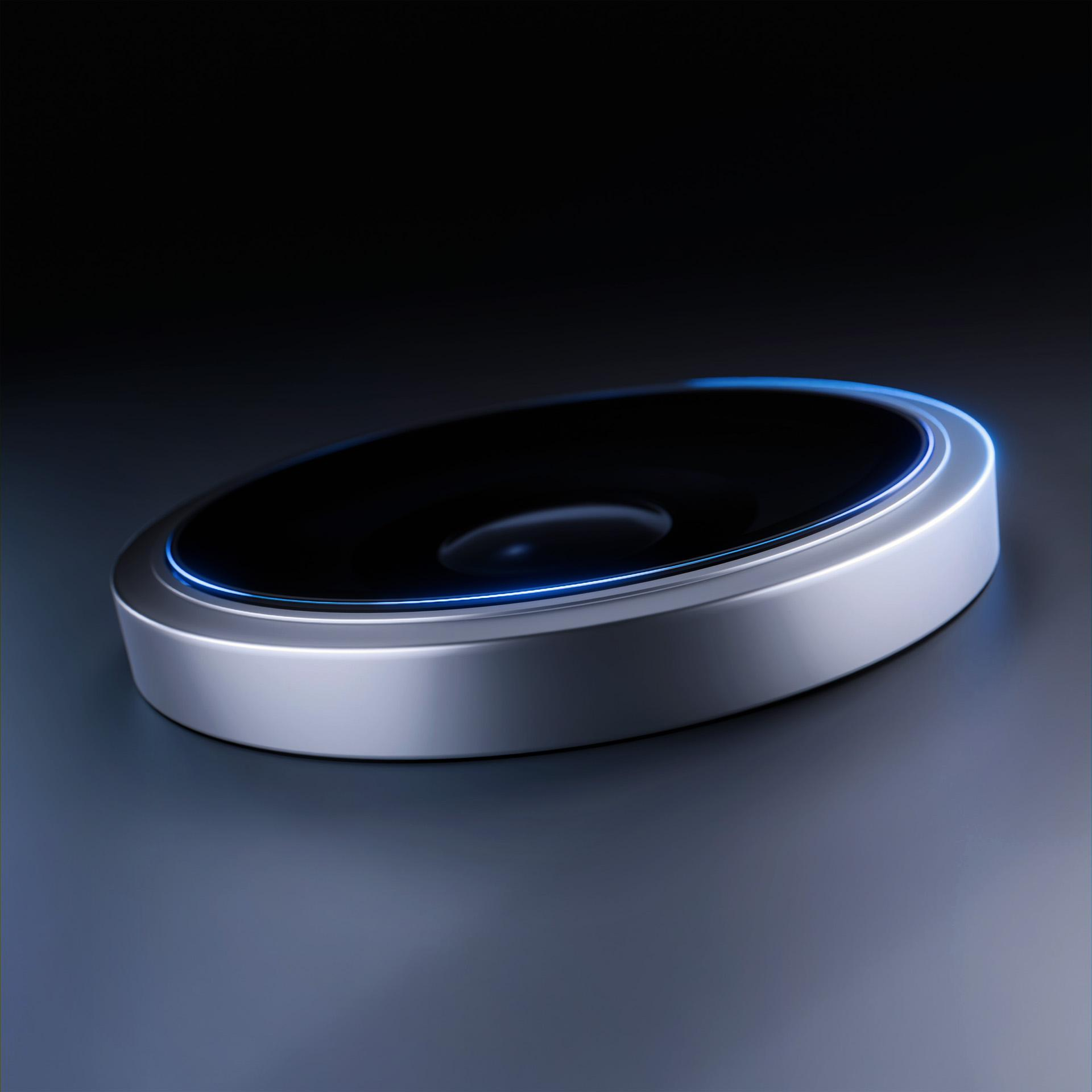 Un dispositivo argentato e circolare con un centro nero e una luce blu sul bordo, su una superficie scura e riflettente.