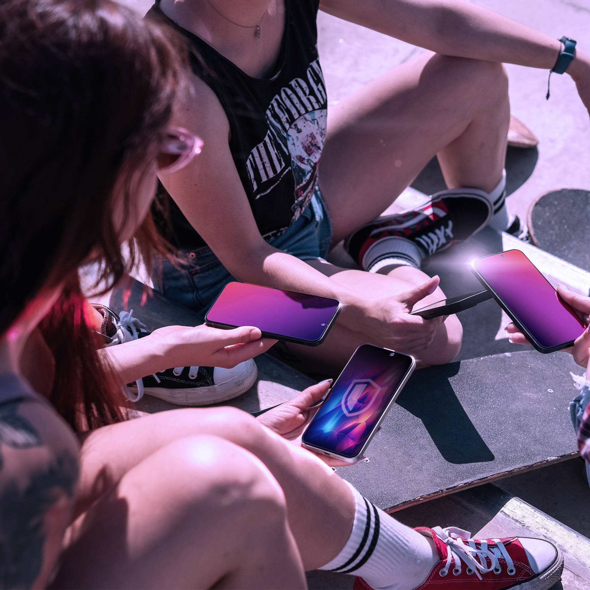 Cztery osoby siedzą ze deskorolkami, trzymając smartfony. Ekrany pokazują gradienty kolorów lub ikonę tarczy.