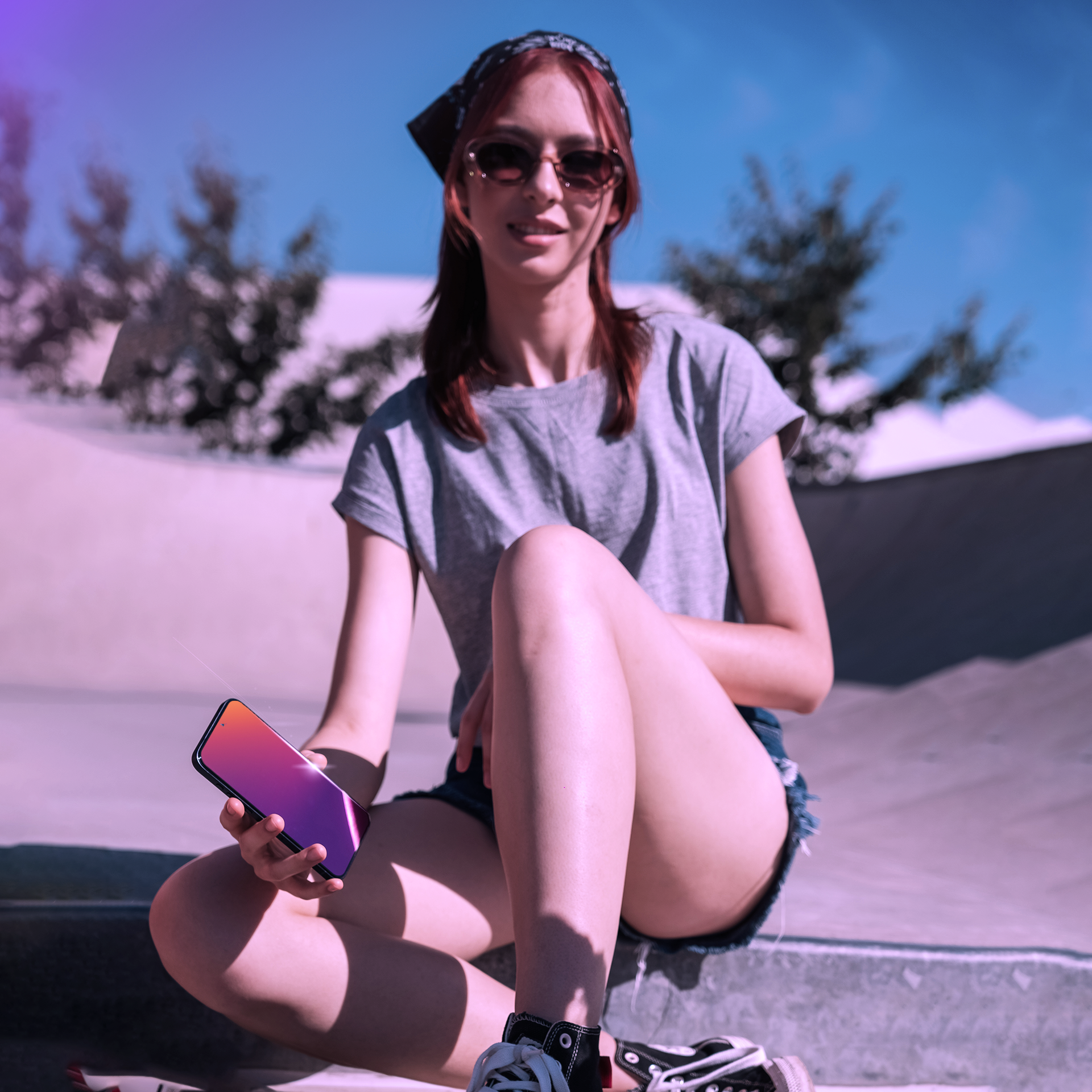 Kobieta w okularach przeciwsłonecznych trzyma telefon. Siedzi na rampie, ubrana w szarą koszulkę, dżinsowe szorty i bandanę.