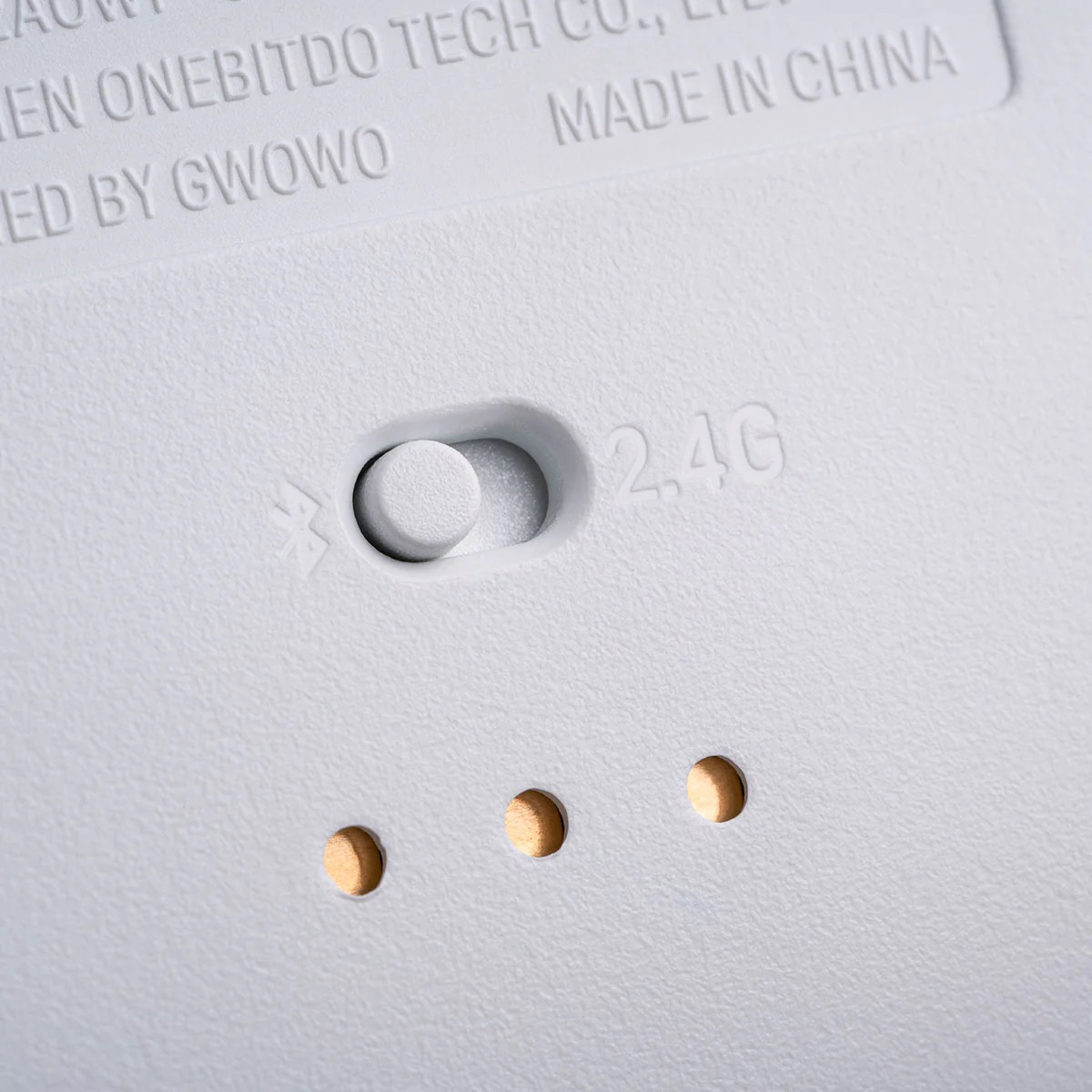 Zbliżenie białego urządzenia z przełącznikiem i trzema małymi otworami. Tekst zawiera "2.4G", "Made in China" i informacje o firmie.