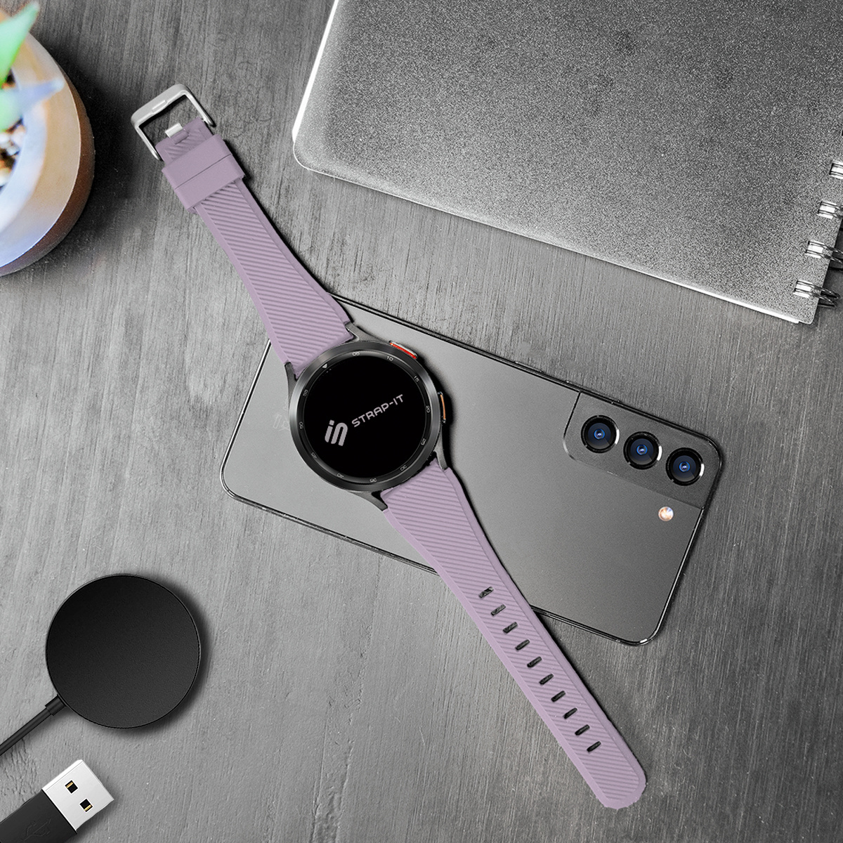 Eine Smartwatch mit lavendelfarbenem Armband liegt auf einem Telefon. Ein kabelloses Ladegerät und ein Notizbuch sind ebenfalls zu sehen.