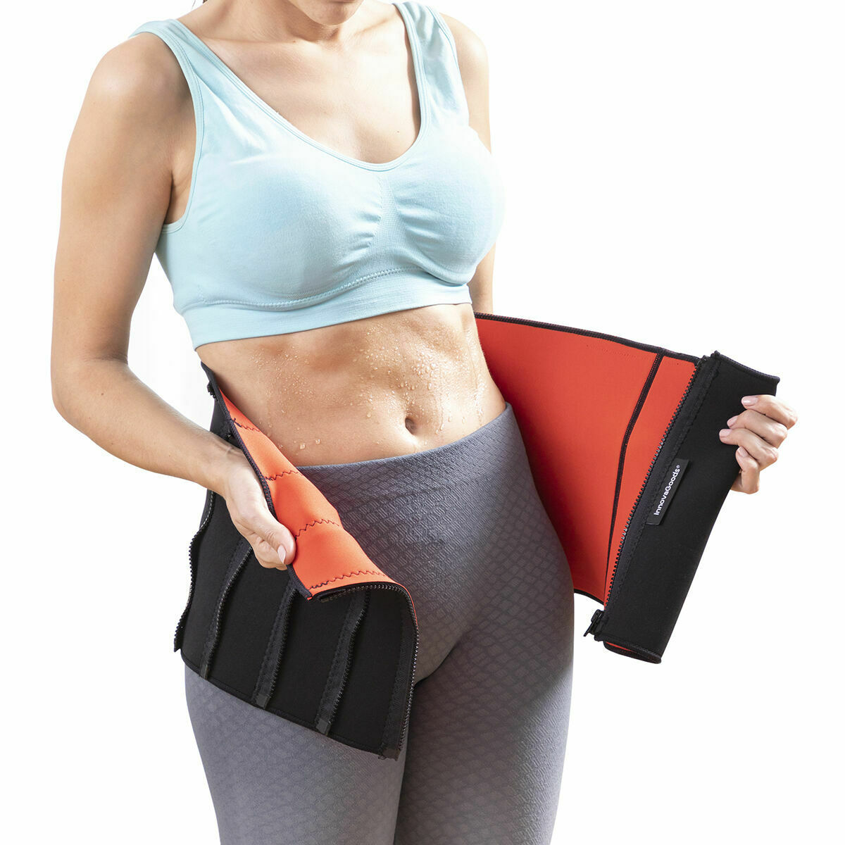 Eine Frau in einem blauen Sport-BH und grauen Leggings zieht einen schwarz-orangenen Taillentrainer an.