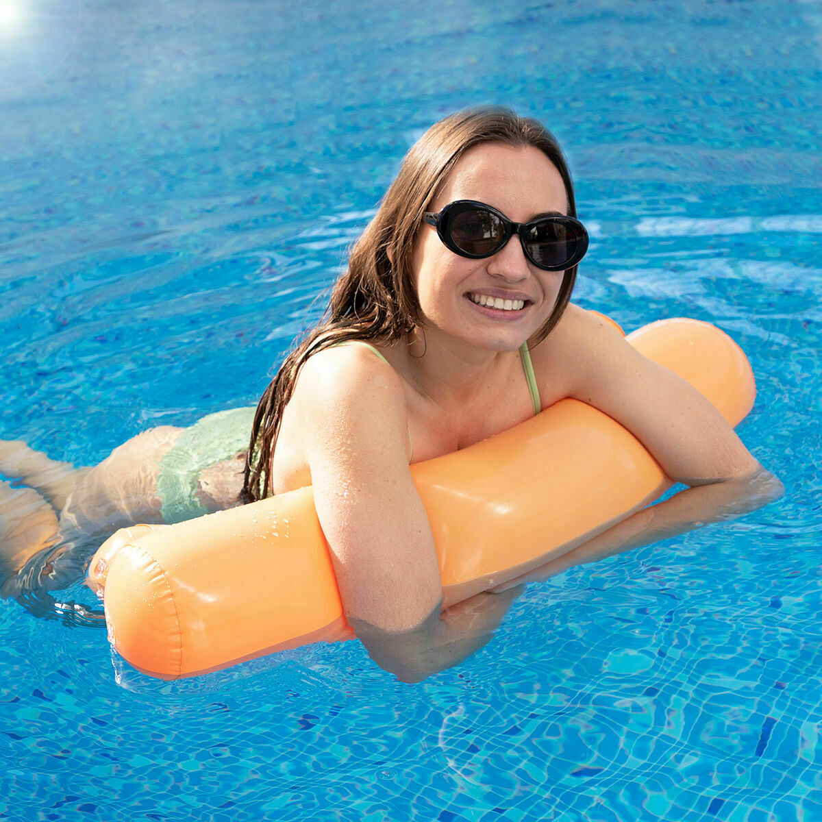 Una mujer con gafas de sol sonríe en una piscina, sosteniendo un inflable naranja. La piscina es azul.