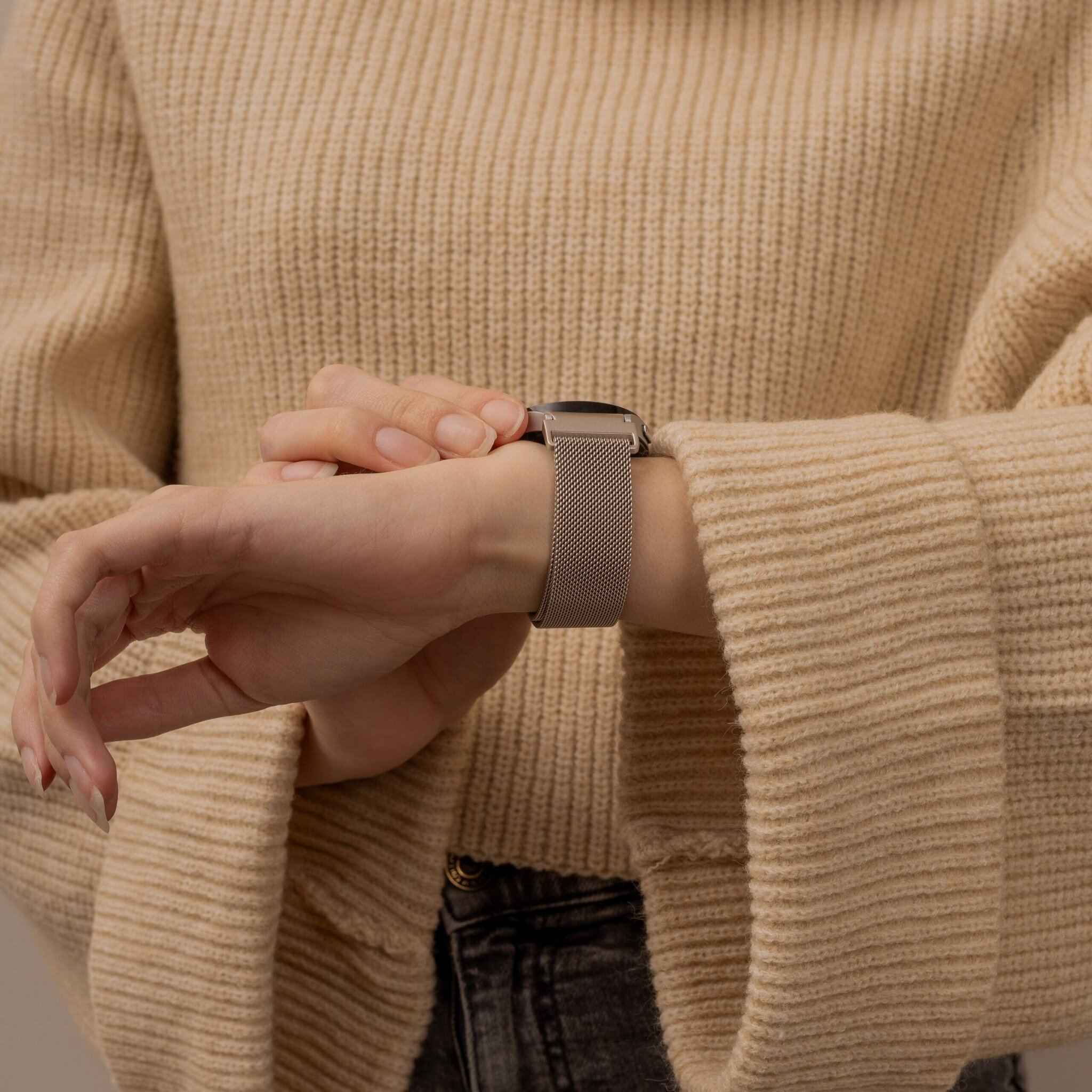 Person trägt einen beigefarbenen Pullover und eine Uhr. Die Uhr hat ein Metallband. Die Person berührt die Uhr.