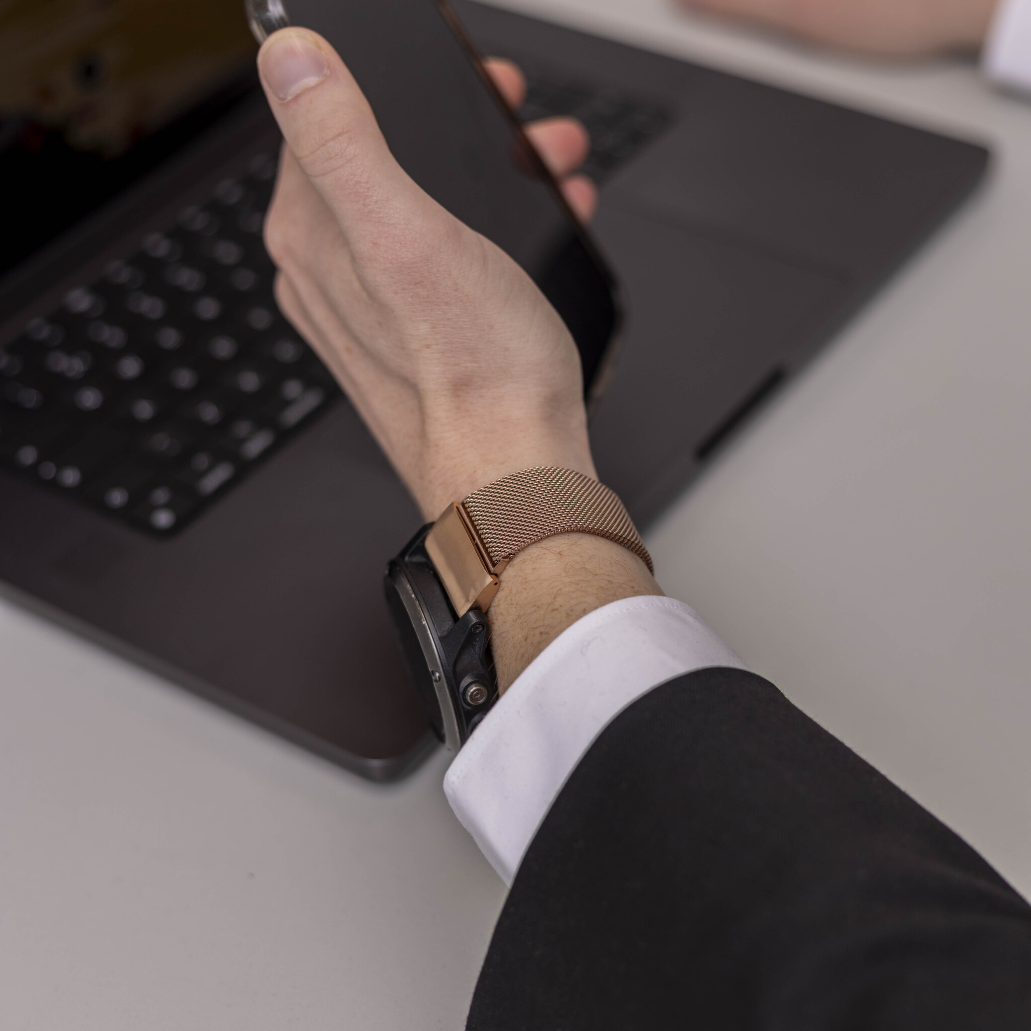 Eine Person hält ein Telefon über einem Laptop. Eine Uhr befindet sich am Handgelenk. Die Person trägt einen schwarzen Anzug und ein weißes Hemd.