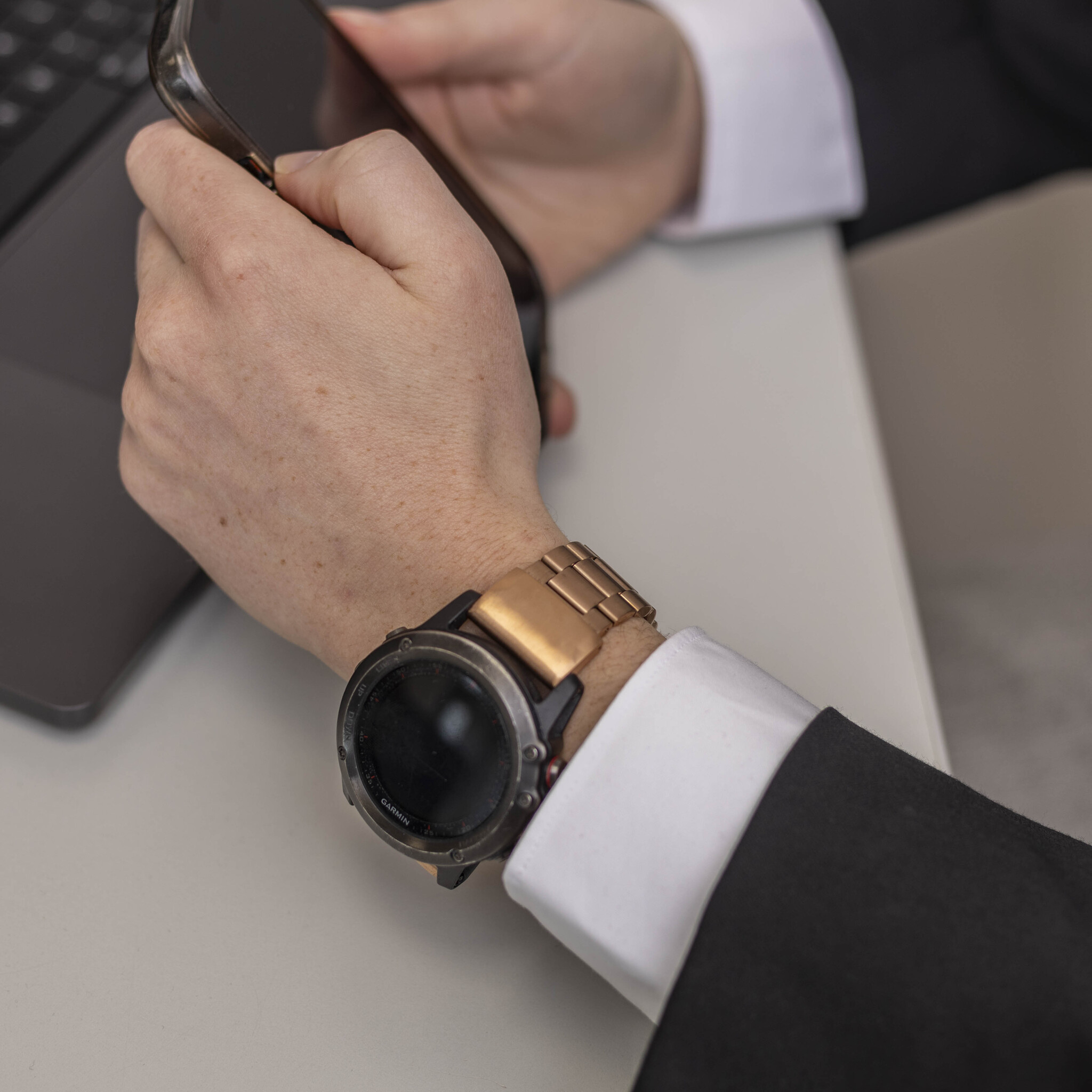 Das Handgelenk einer Person mit goldener Uhr hält ein Telefon. Ein schwarzer Anzug und ein weißes Hemd sind ebenfalls zu sehen.