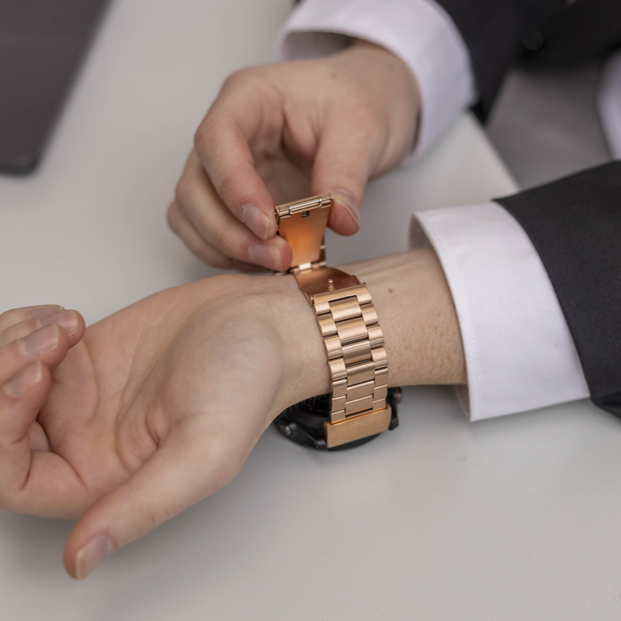 Eine Person befestigt eine goldene Uhr. Die Uhr befindet sich am Handgelenk, neben einer Hemdmanschette. Der Hintergrund ist weiß.