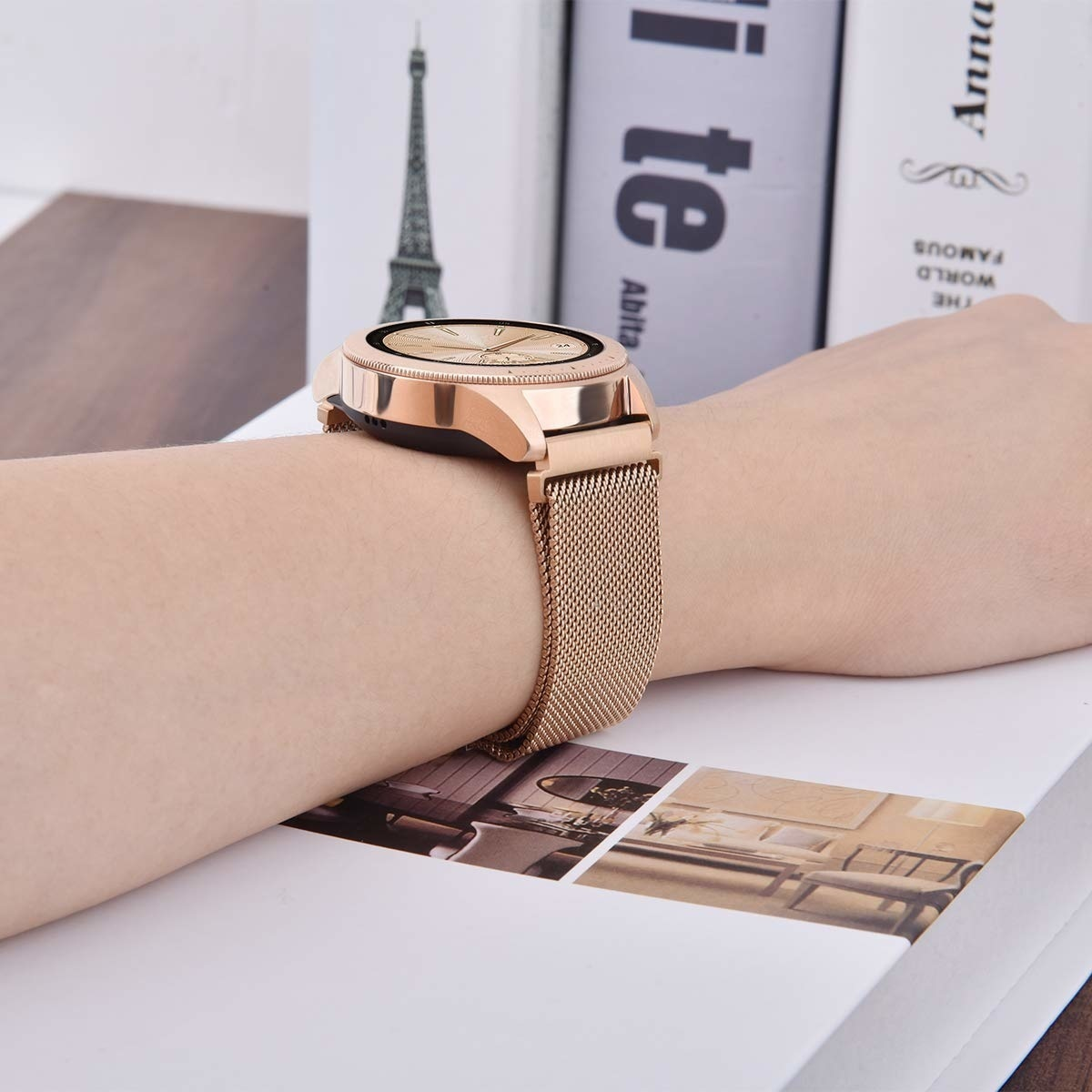 Eine goldene Smartwatch befindet sich an einem Handgelenk mit einem Mesh-Armband. Sie liegt auf einem weißen Buch, im Hintergrund ein Eiffelturm.