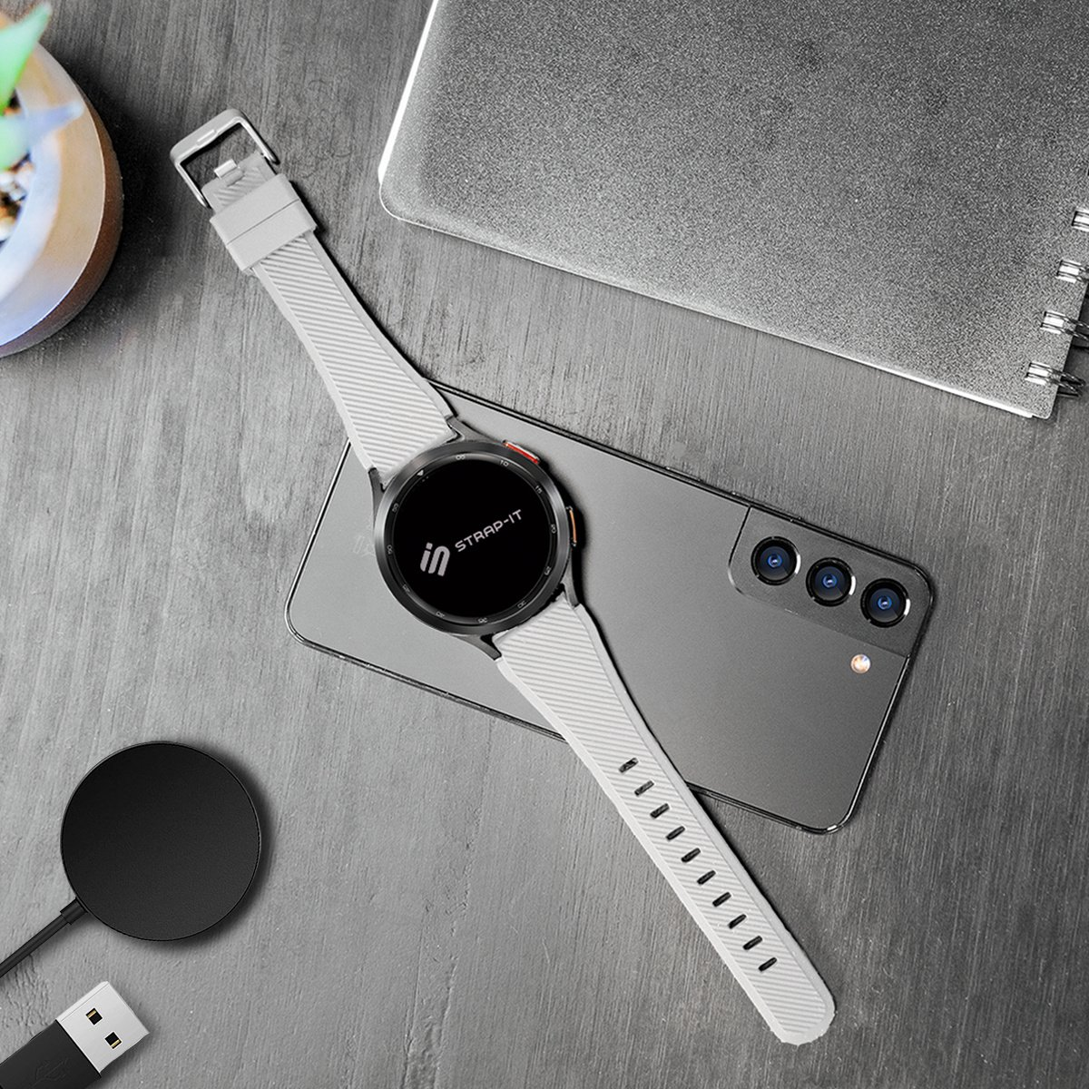 Eine graue Smartwatch mit weißem Armband liegt auf einem grauen Telefon. Ein schwarzes Ladegerät und ein Notizbuch sind in der Nähe.