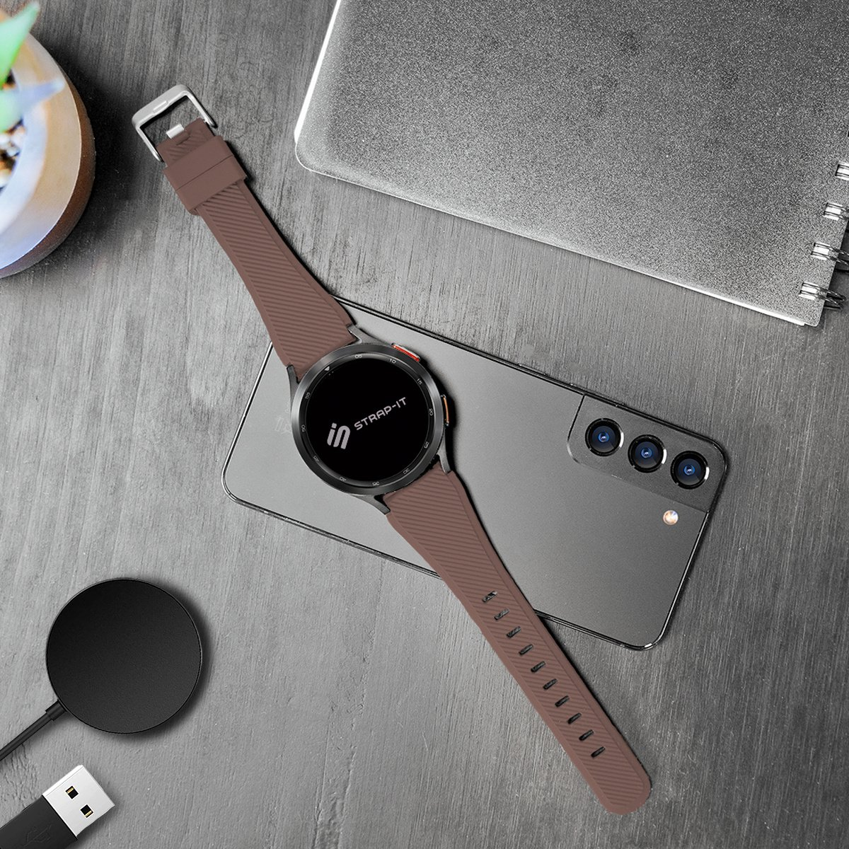 Eine braune Smartwatch mit Armband liegt auf einem Smartphone. Ein Ladegerät und ein Notizbuch liegen daneben. Die Uhr trägt die Marke 'strap-it'.
