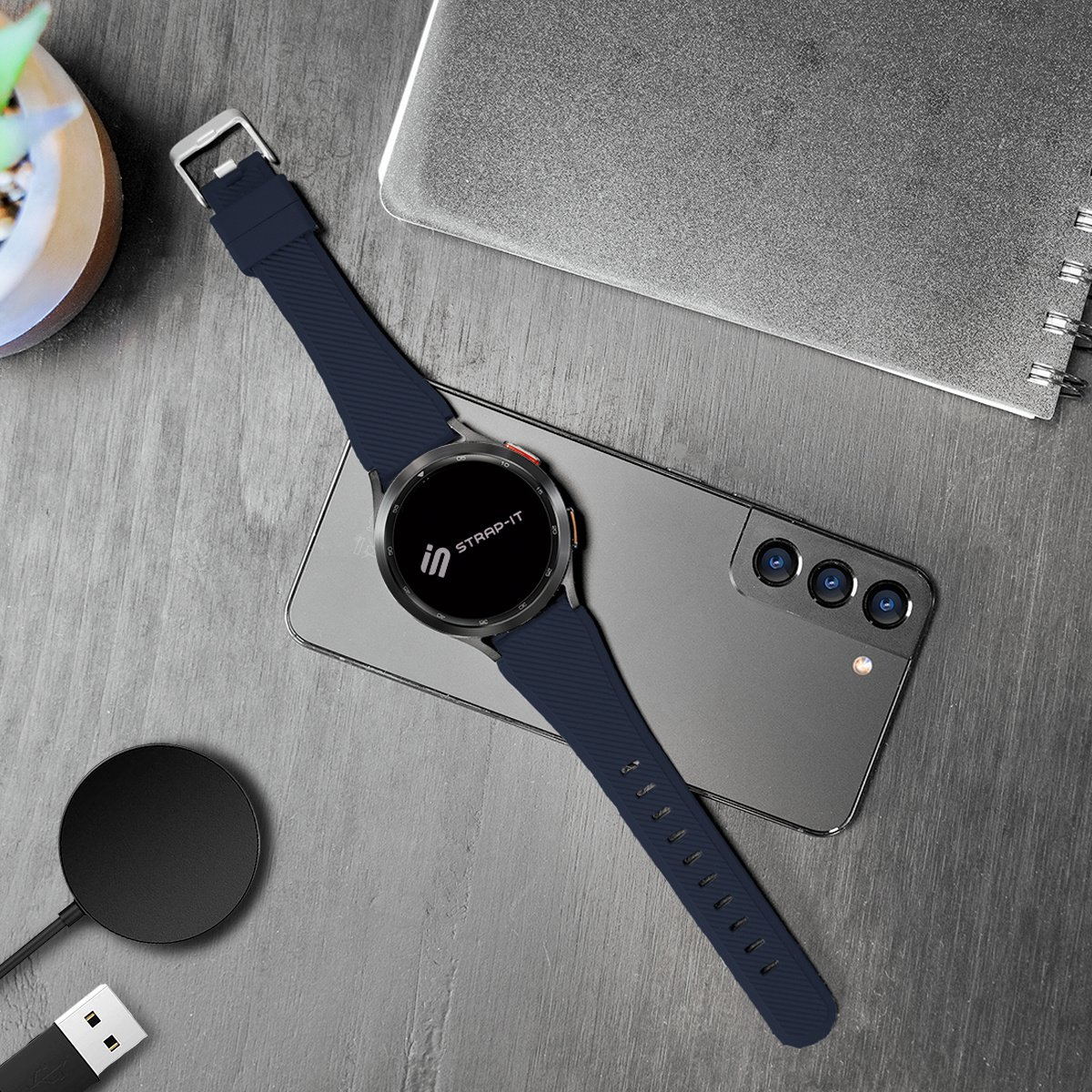 Eine marineblaue Smartwatch mit passendem Armband liegt auf einem Telefon. Ein Ladegerät, ein Notizbuch und eine kleine Pflanze sind ebenfalls vorhanden.