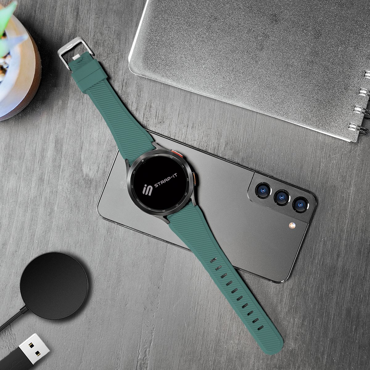 Eine Smartwatch mit tealfarbenem Armband liegt auf einem Telefon. Ein Ladepad und ein Notizbuch befinden sich ebenfalls auf der grauen Oberfläche.