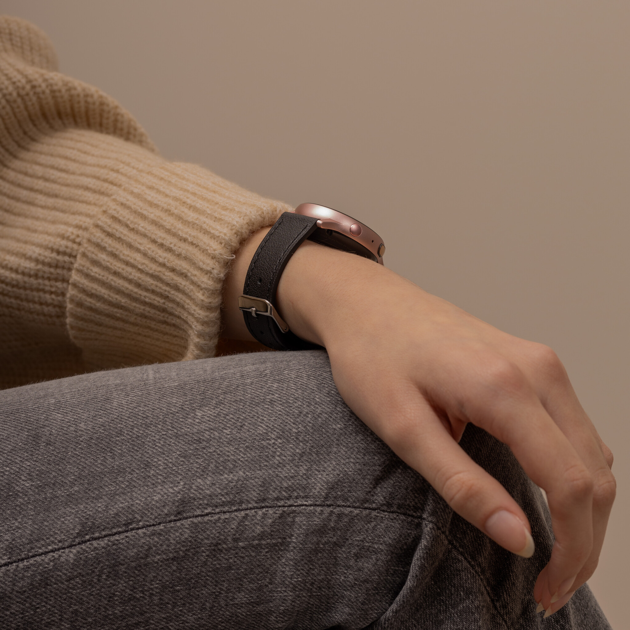 Ein Arm mit roségoldener Uhr an schwarzem Lederarmband, auf grauer Jeans liegend.