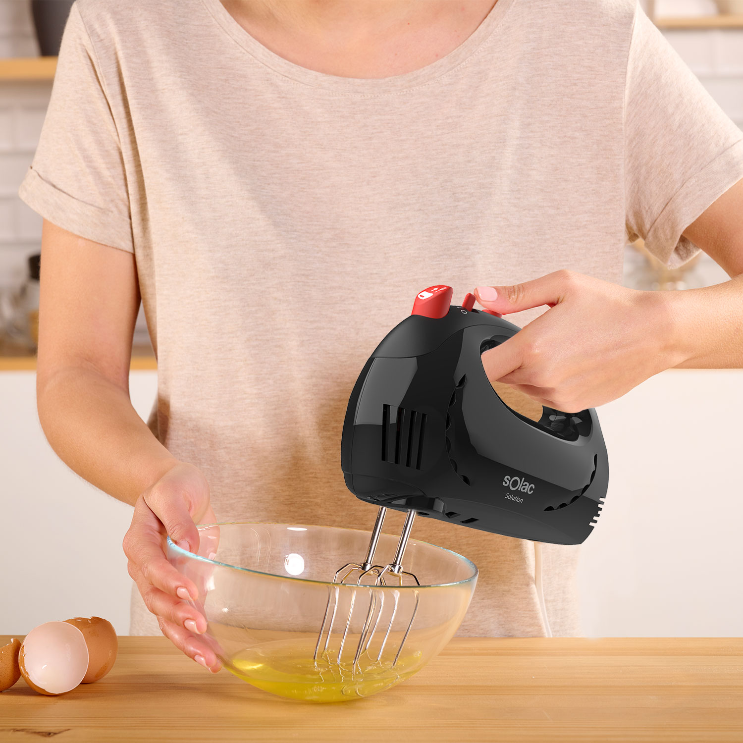 Eine Frau verquirlt mit einem schwarzen Solac-Handmixer Eiweiß in einer Glasschüssel. Eierschalen liegen auf einer Holzoberfläche.