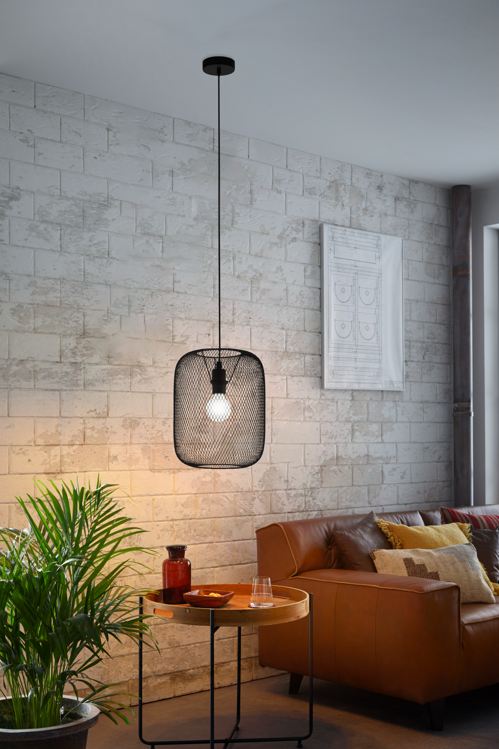 Eine schwarze Pendelleuchte hängt über einem kleinen Tisch mit Dekoration. Ein Ledersofa und eine Ziegelmauer sind im Hintergrund.