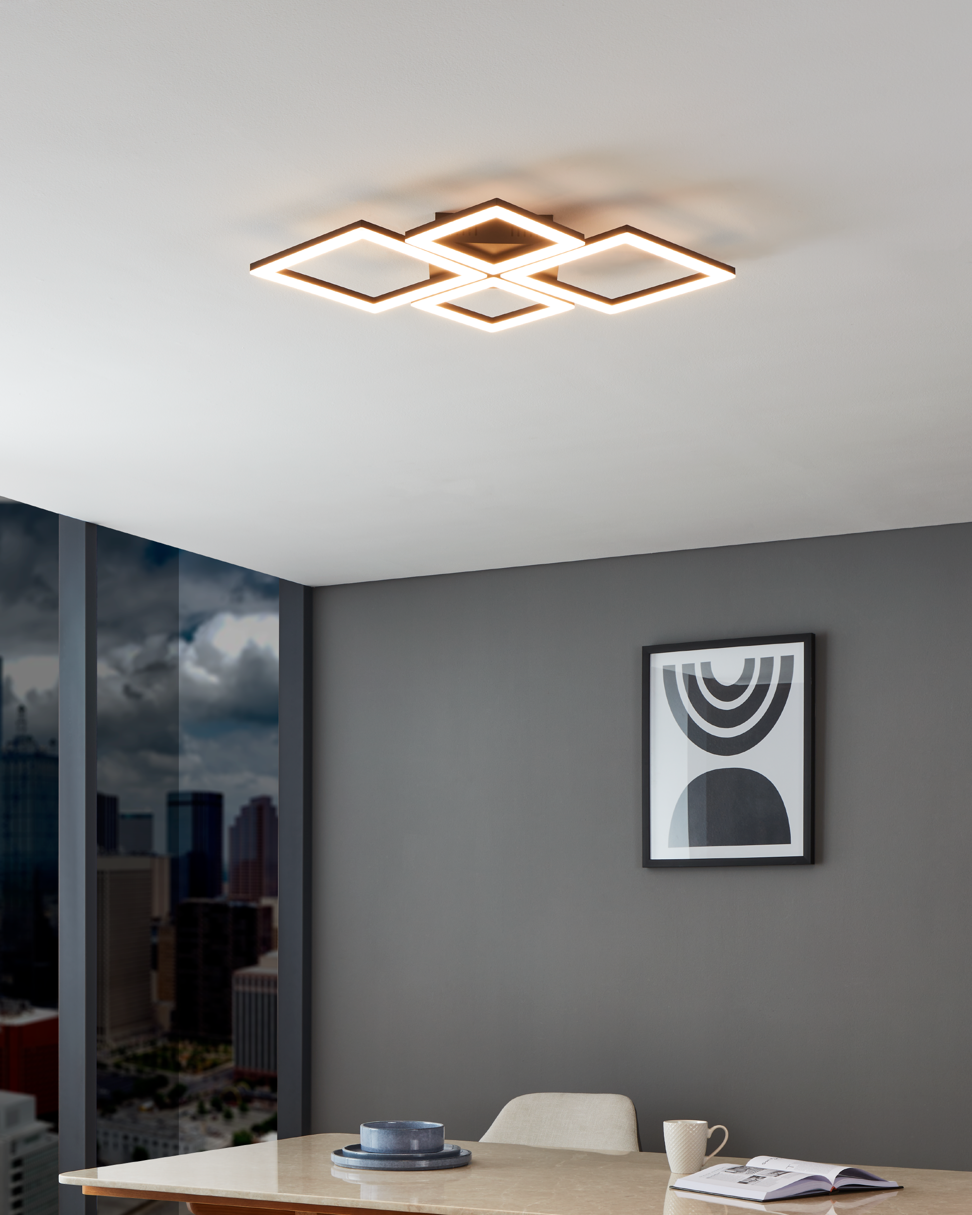 Una stanza moderna con una luce a soffitto geometrica. Un tavolo apparecchiato con ciotola, piatti e libro. Vista della città.