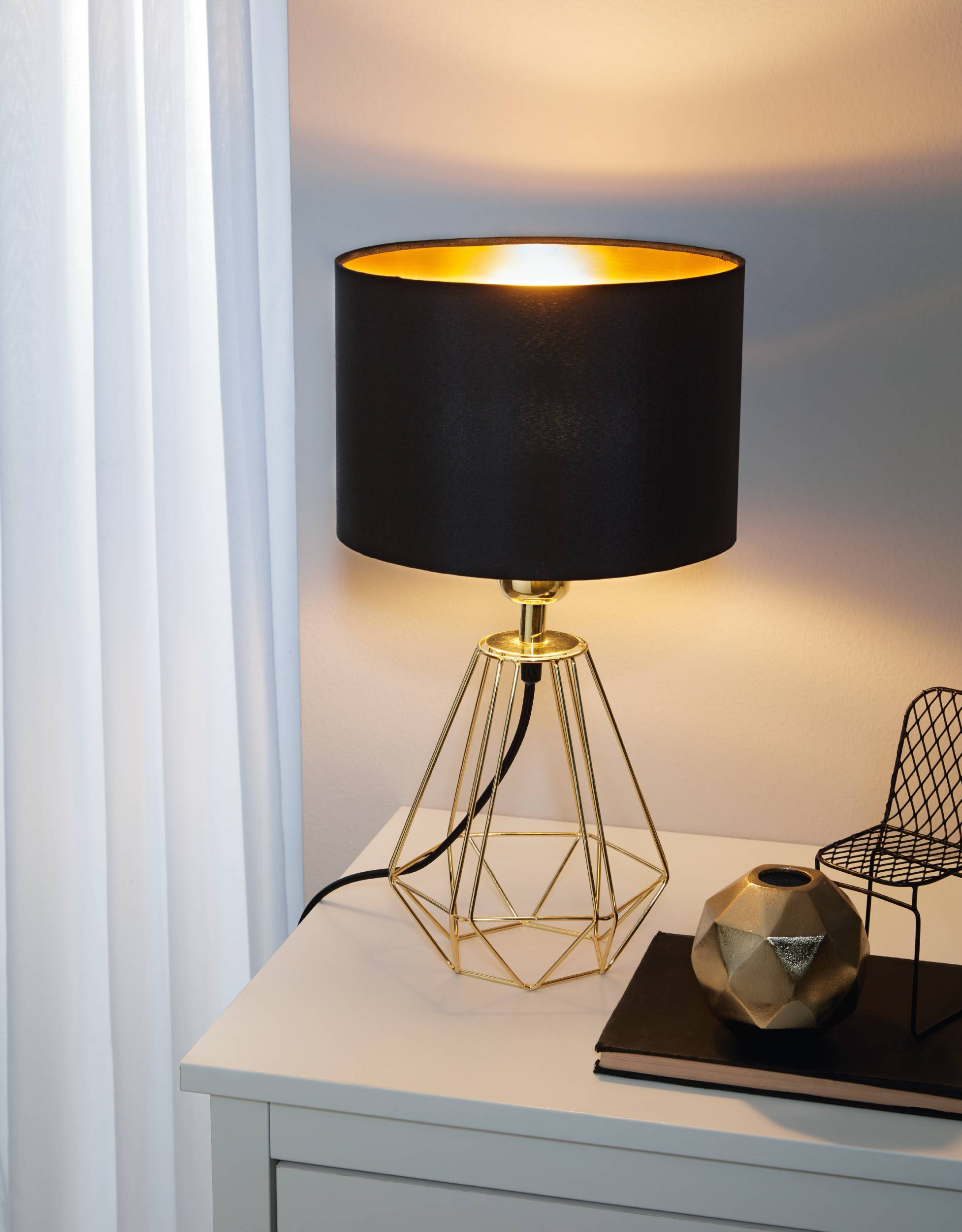 Un paralume nero è posto su una lampada geometrica dorata su un tavolo bianco. Un vaso d'oro e una sedia nera sono anche sul tavolo.