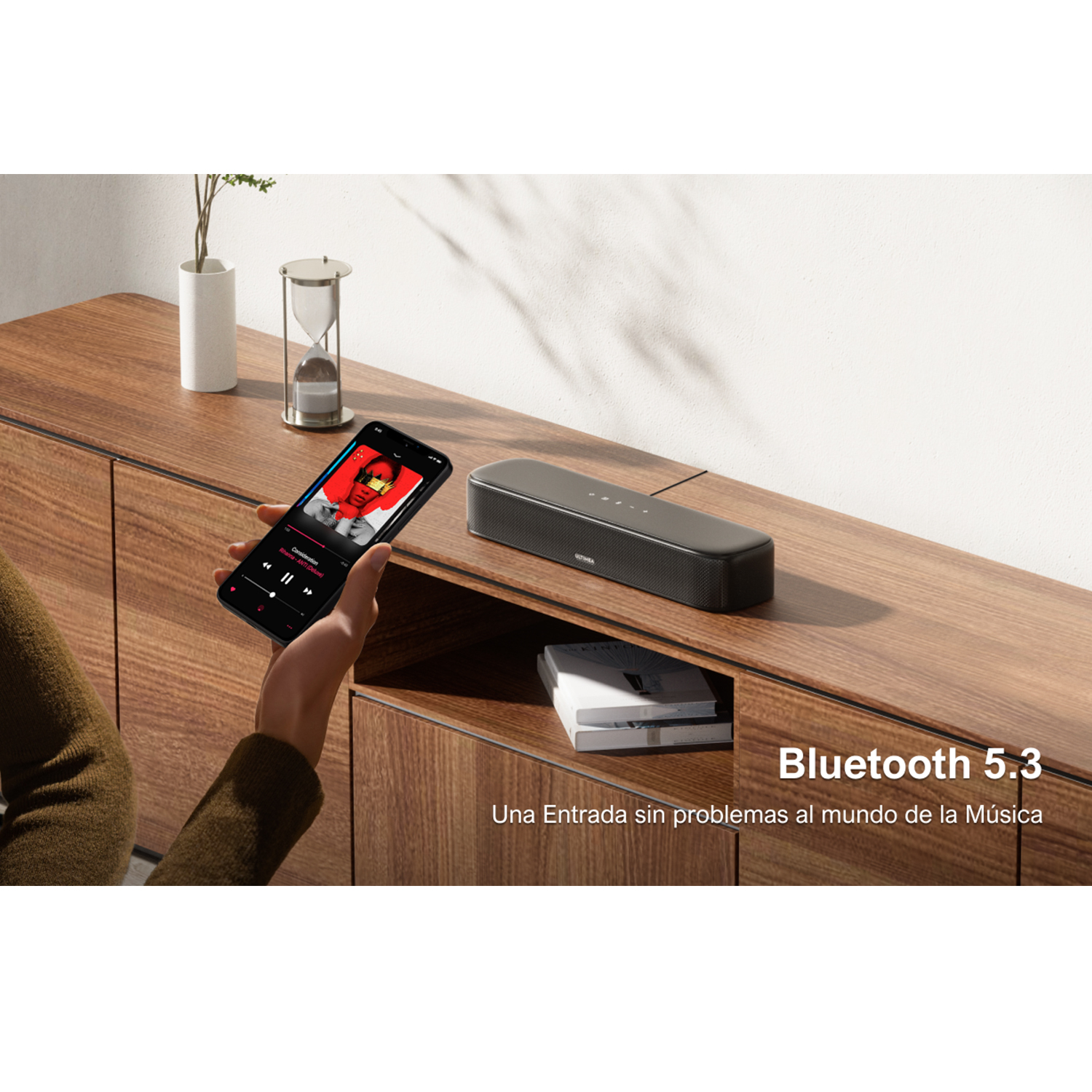 Una mano sostiene un teléfono reproduciendo música, junto a un altavoz. Un mueble de madera está en el fondo.