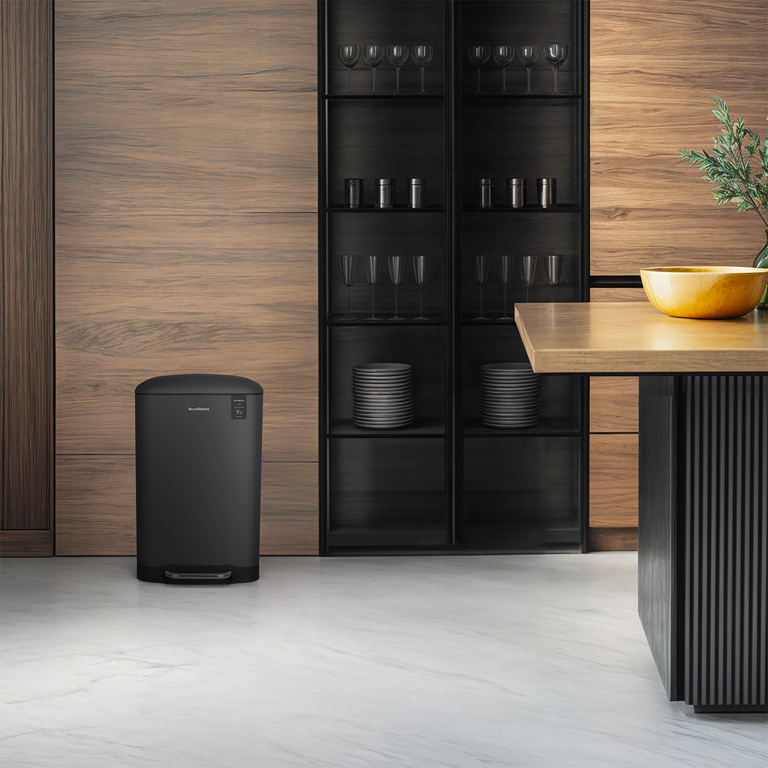 Contenedor de basura negro en una cocina moderna. Gabinetes de madera, estantes de vidrio con platos y vasos. Piso de mármol y mesa de madera.