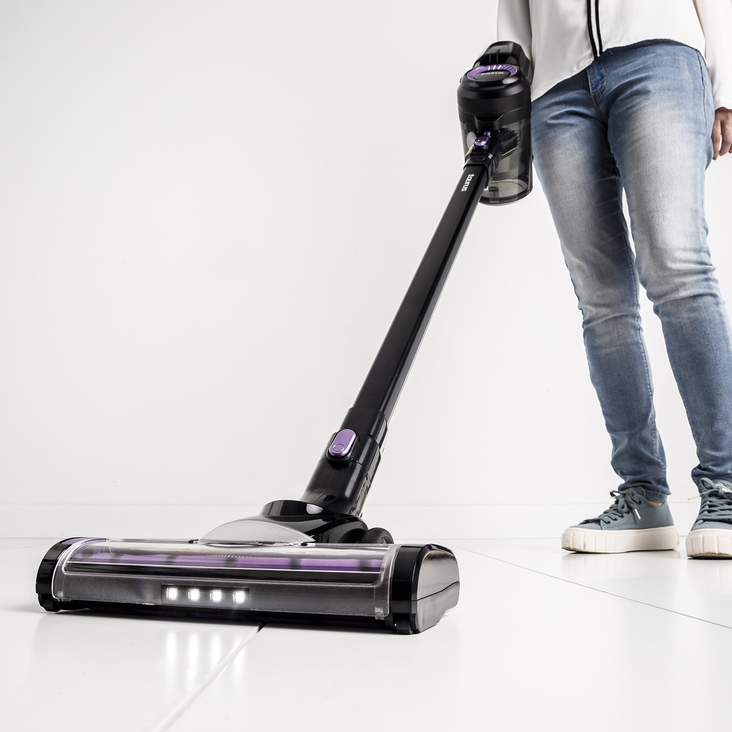 Persona che usa un aspirapolvere nero e viola su un pavimento bianco. La persona indossa jeans e scarpe da ginnastica. Lo sfondo è bianco.