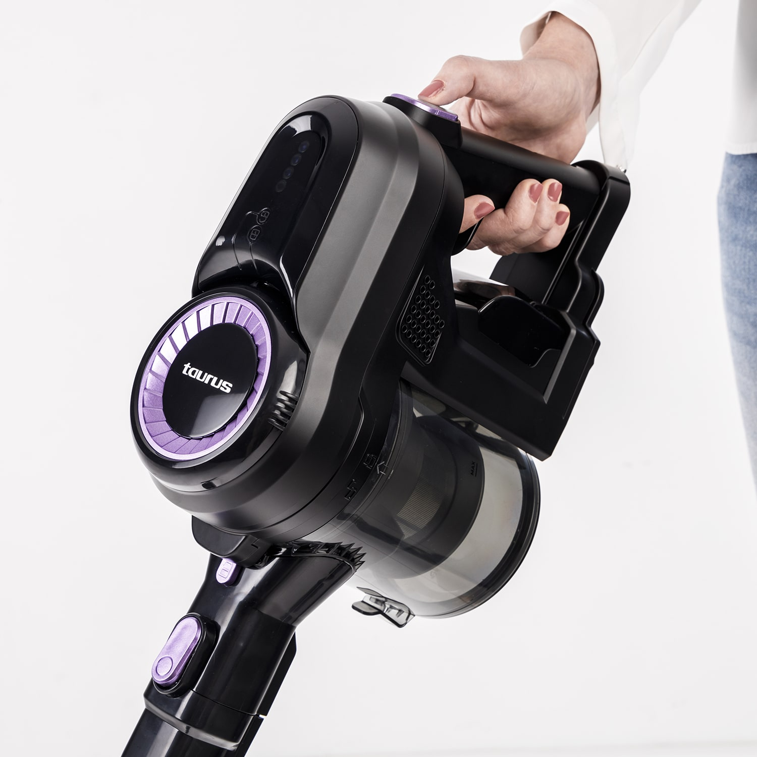 Una persona tiene in mano un aspirapolvere Taurus nero e viola. L'aspirapolvere ha un contenitore per la polvere trasparente e si trova su sfondo bianco.