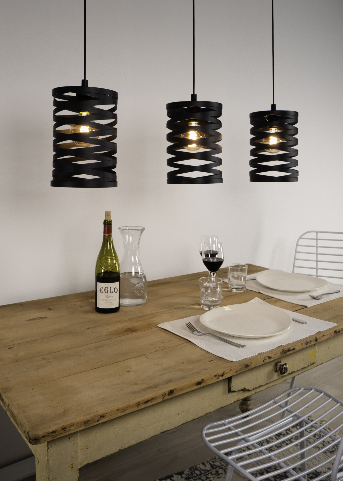 Eine Szene mit drei schwarzen Lampen, einem Holztisch, Wein, Tellern und Gläsern. Im Hintergrund ein Metallstuhl.