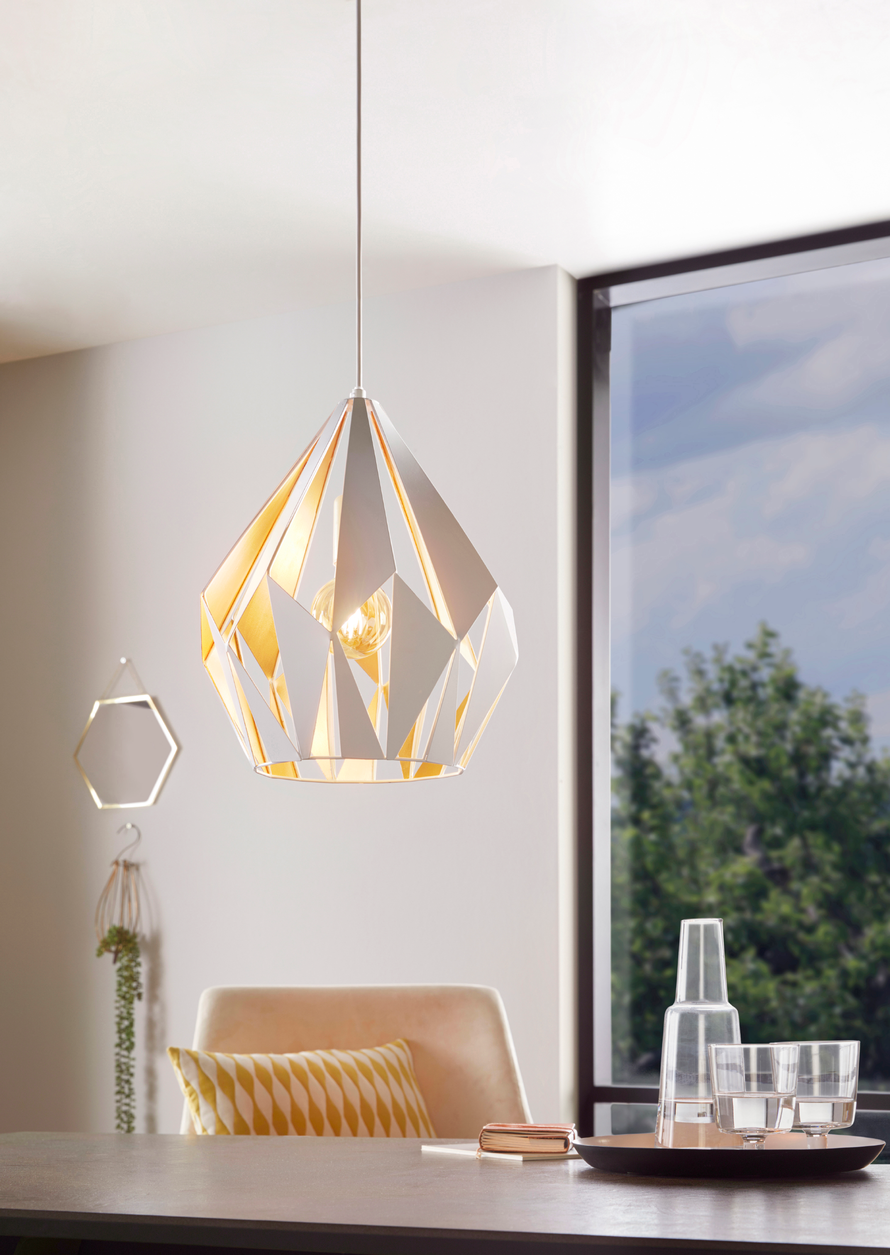 Un lampadario moderno è appeso sopra un tavolo per due. Una finestra mostra uno sfondo verde e frondoso.