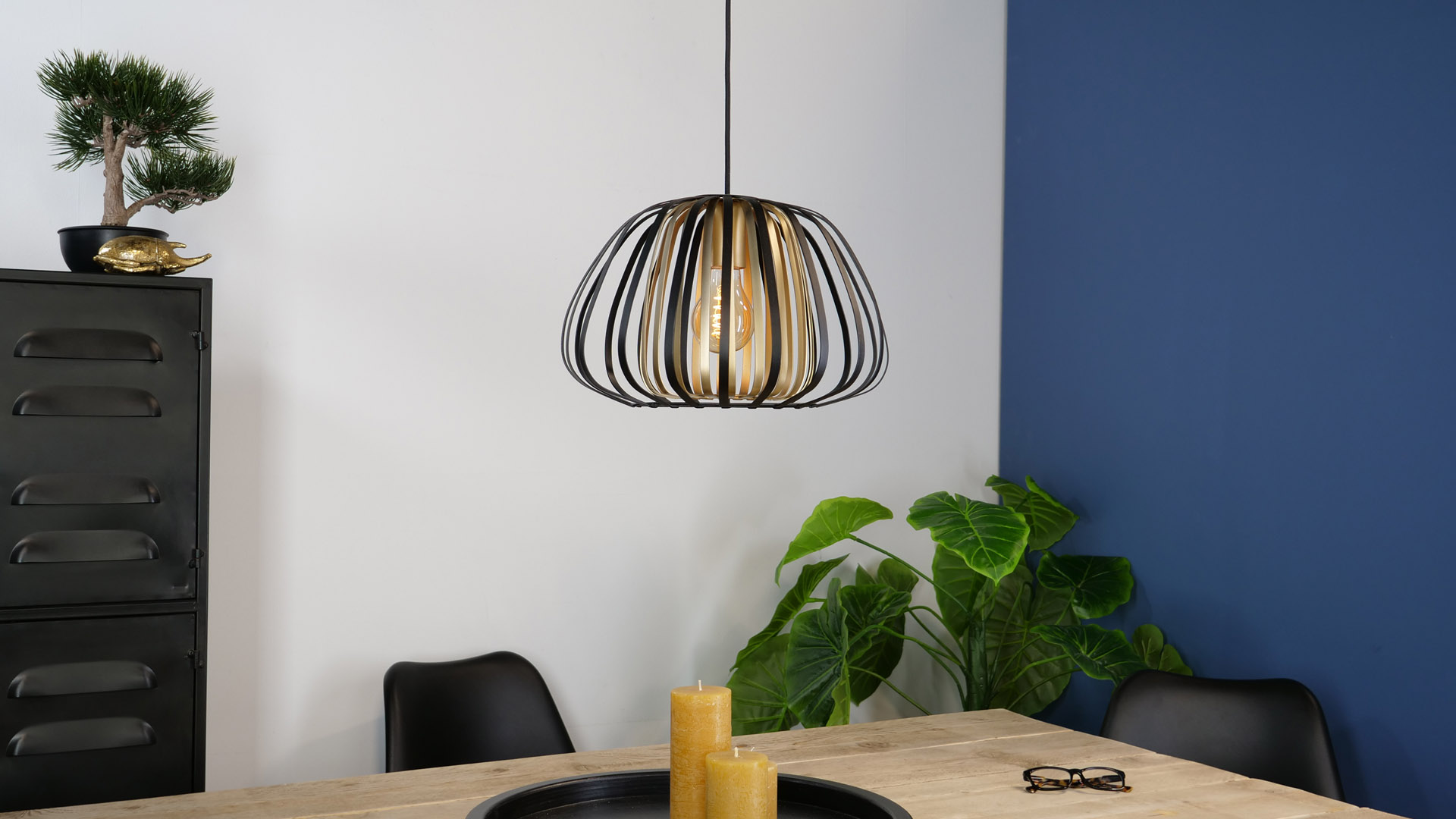 Ein modernes Esszimmer mit Hängelampe, Holztisch, Kerzen und Pflanze. Schwarze Stühle und eine blaue Wand sind sichtbar.