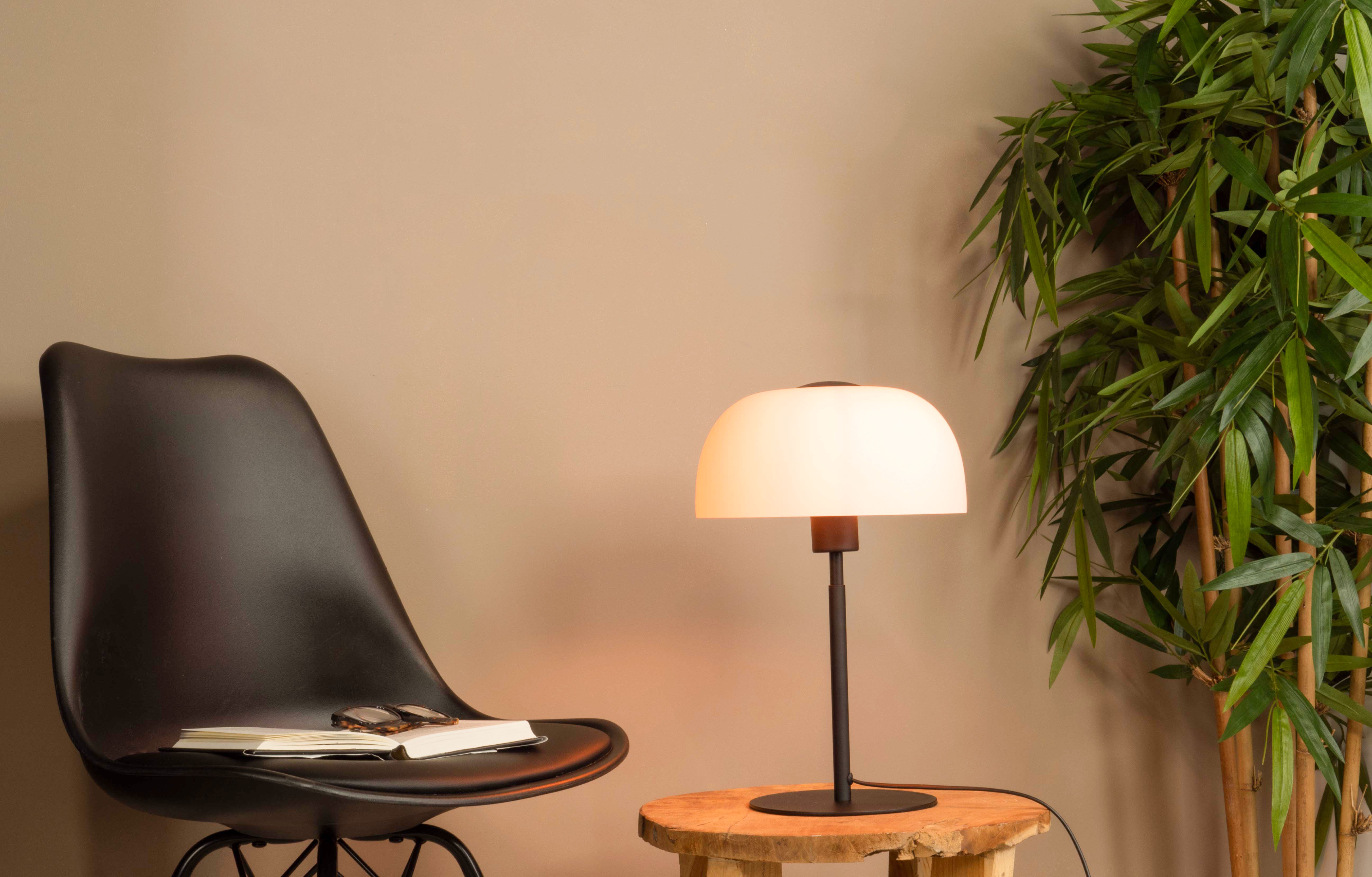 Ein schwarzer Stuhl mit Buch und Brille, eine Lampe auf einem Holztisch und Bambus im Hintergrund.