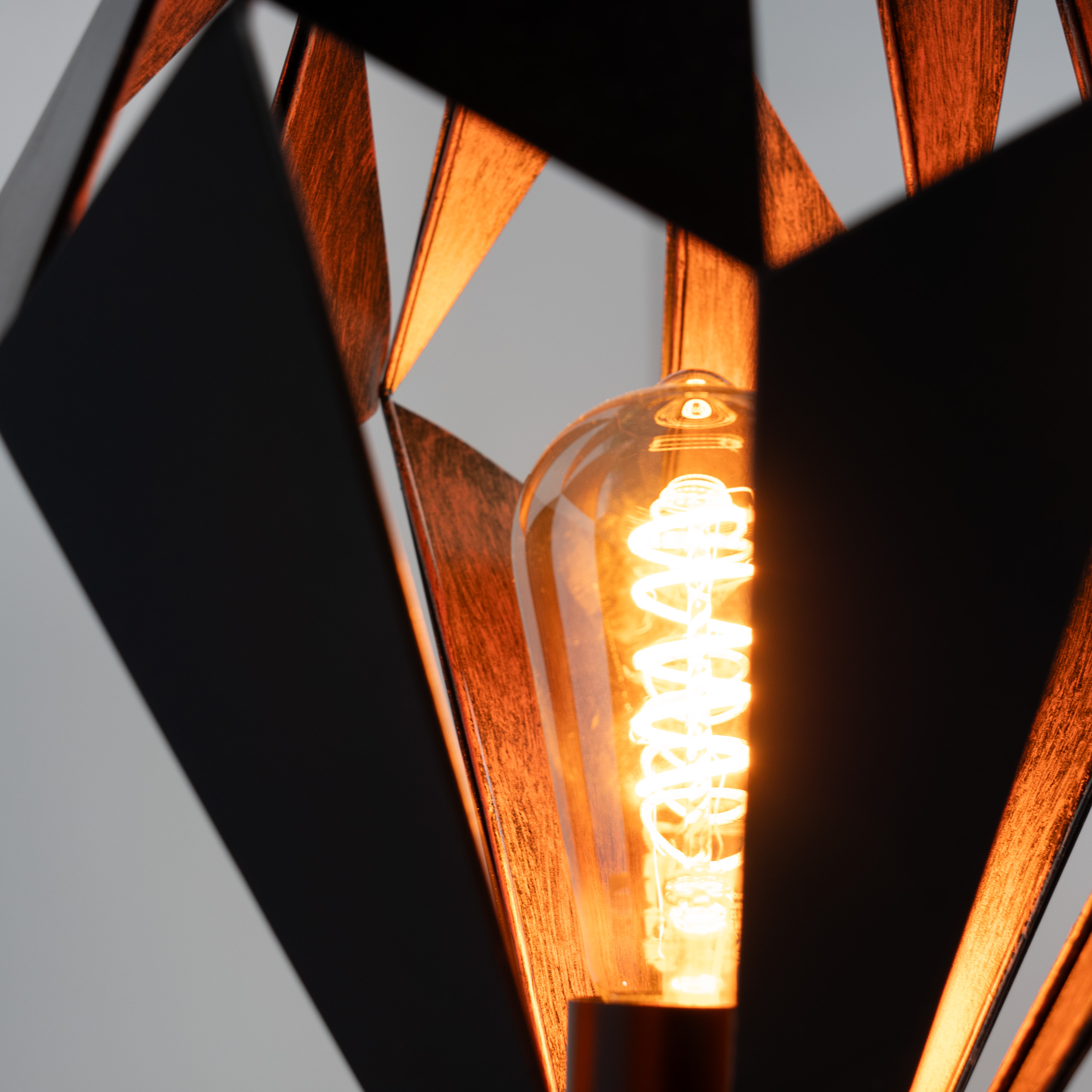Primo piano di una lampada decorativa. Una lampadina a spirale illuminata è incorniciata da una struttura geometrica, scura e metallica.
