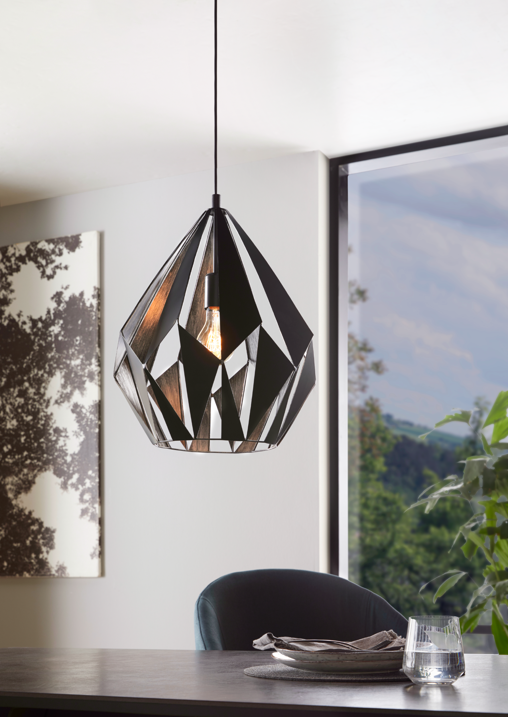 Una lampada a sospensione geometrica è appesa sopra un tavolo apparecchiato per un pasto. Una finestra mostra un paesaggio. C'è un dipinto sul muro.