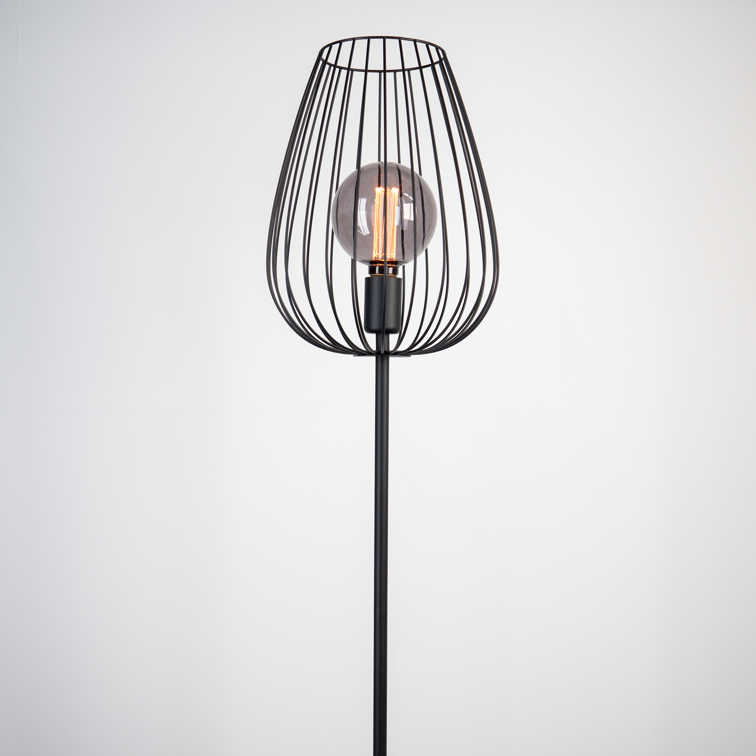 Eine schwarze Lampe mit käfigartigem Schirm, die eine Glühbirne hält. Der Hintergrund ist eine weiße Wand.