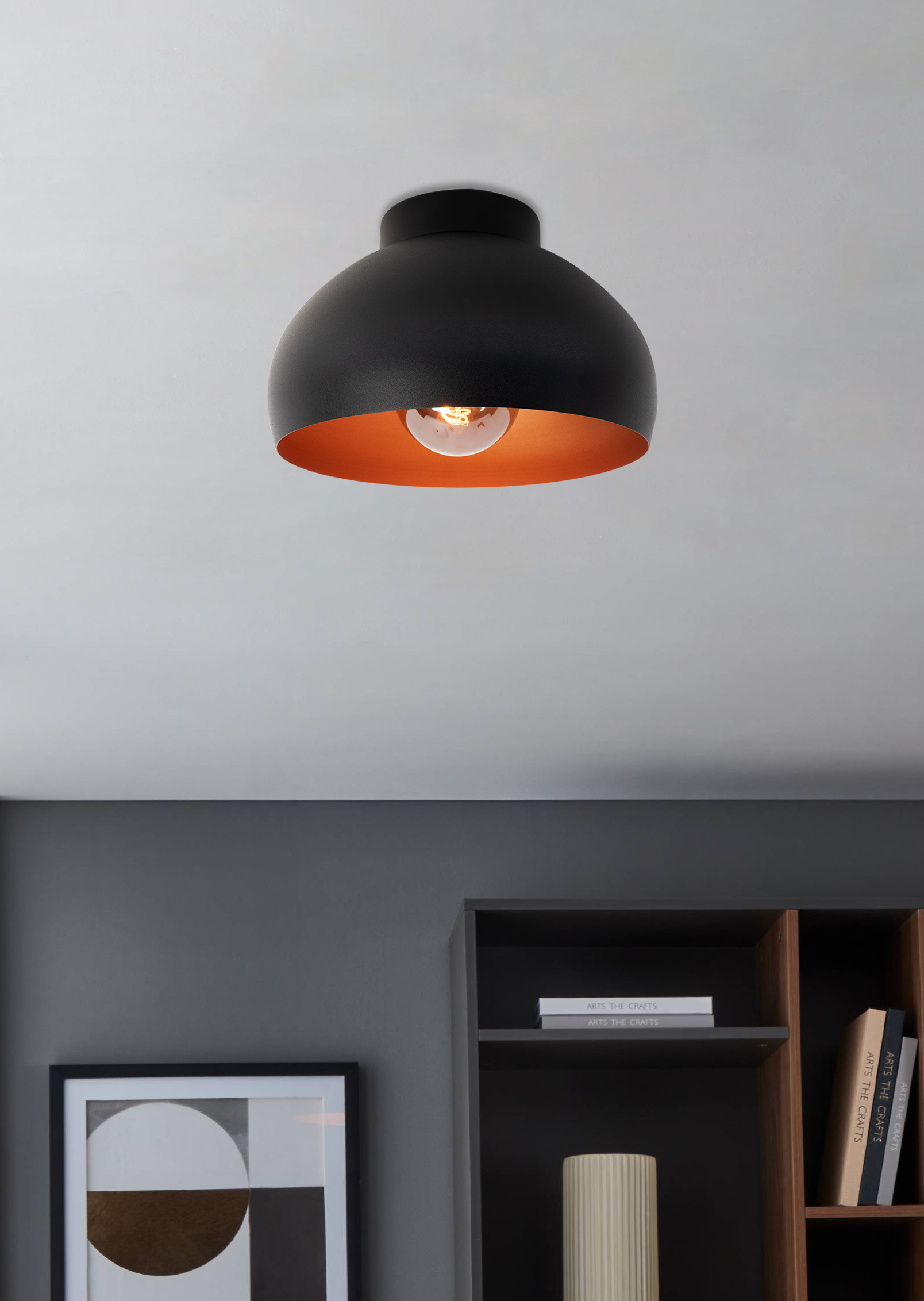 Una lampada da soffitto nera e color rame è appesa a un soffitto bianco. Sotto sono visibili una libreria e un'opera d'arte incorniciata.