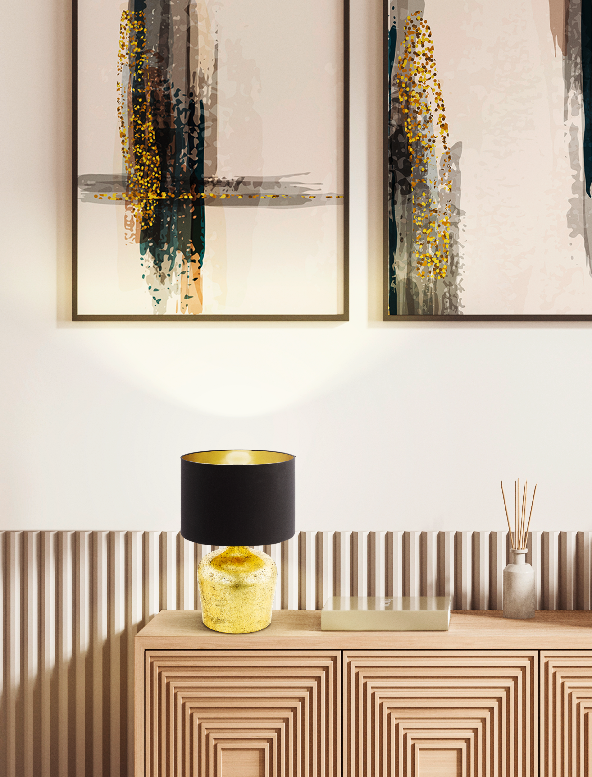 Una lampada con paralume nero e base dorata su un mobile in legno. Dipinti e un vaso sullo sfondo.