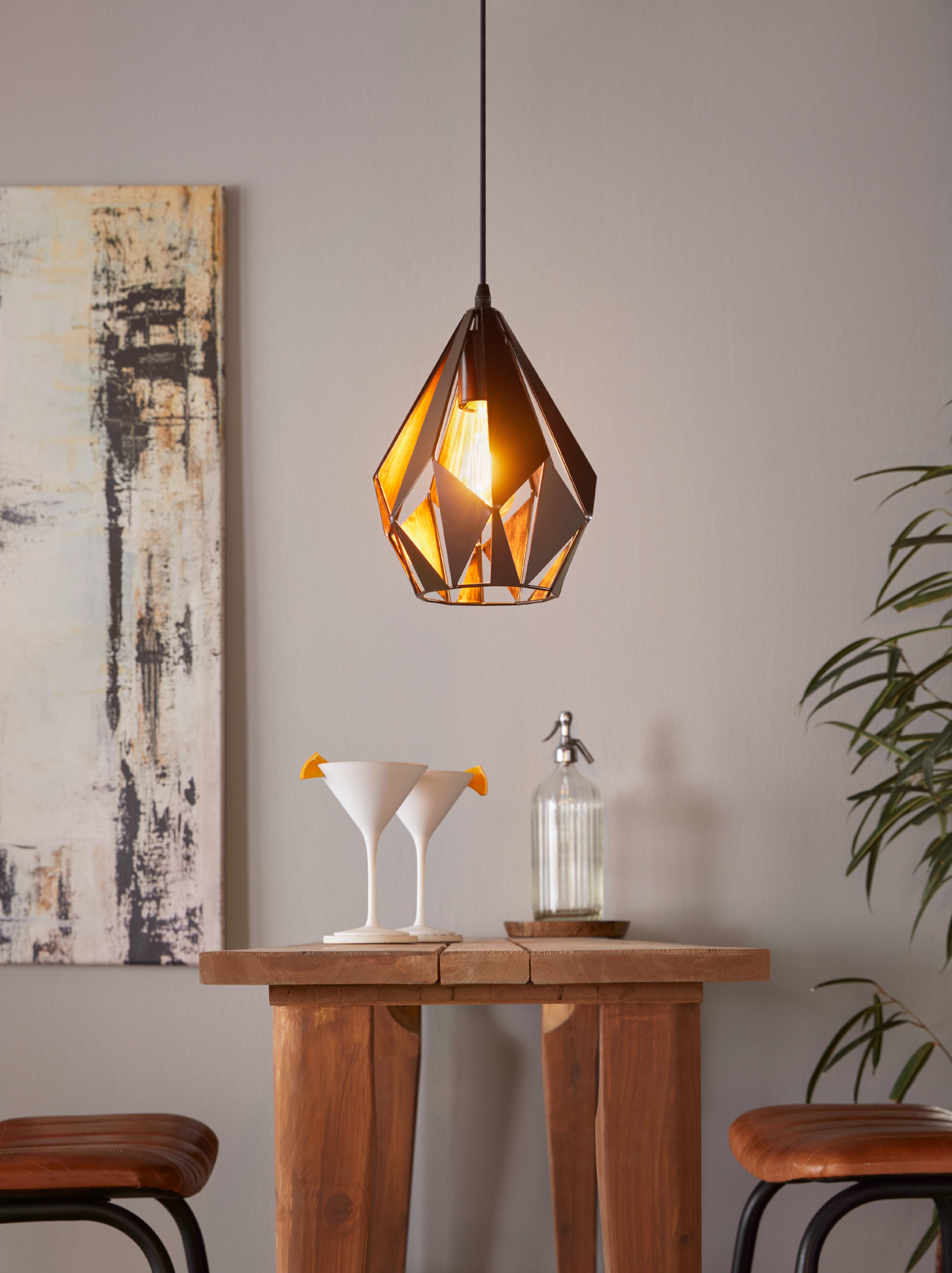 Un ambiente bar moderno con lampada a sospensione geometrica, bicchieri da cocktail e tavolo in legno. Include un dipinto e piante.
