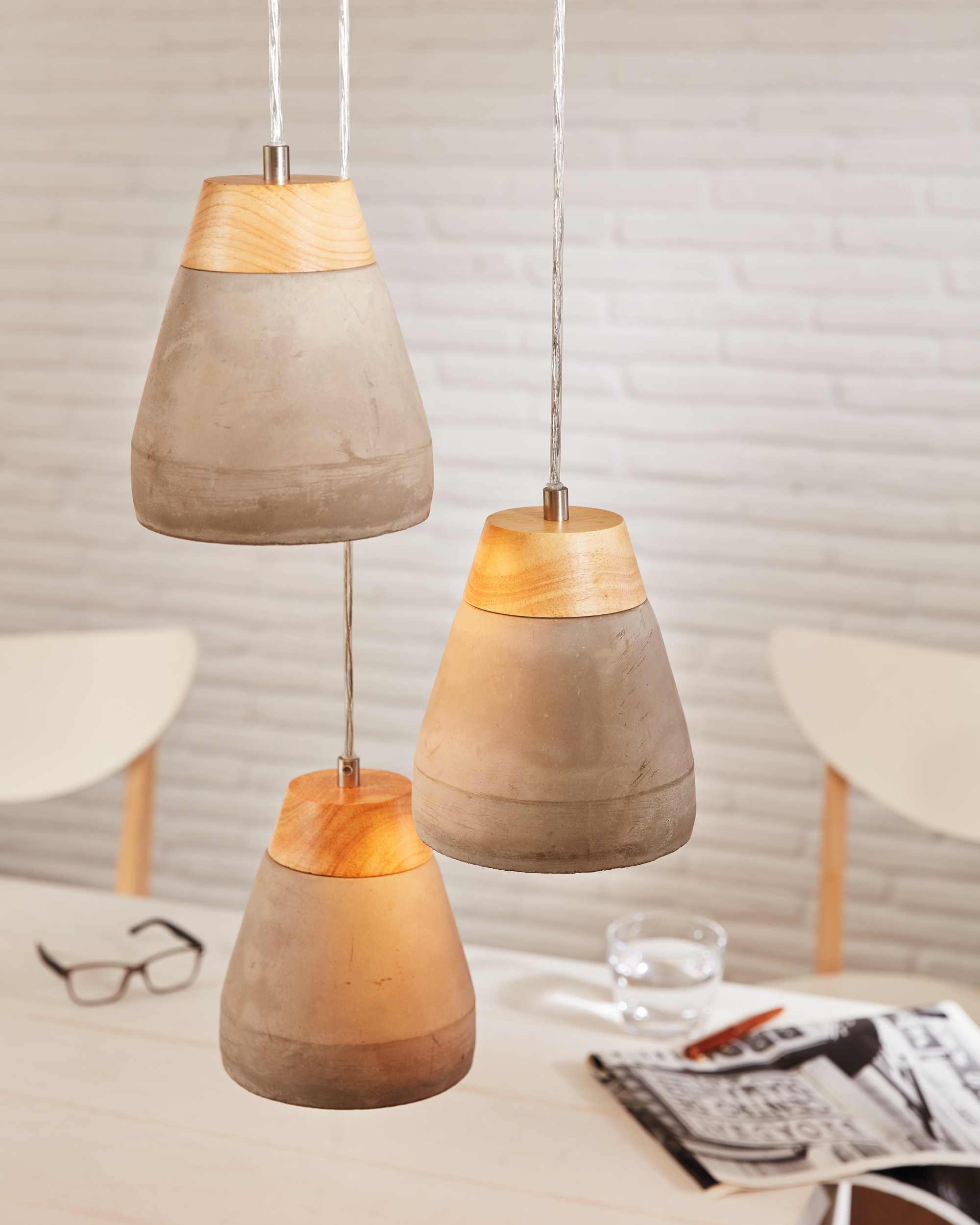 Drei Pendelleuchten mit Betonschirmen und Holzaufsätzen hängen über einem Tisch mit Gläsern, einer Zeitschrift und einem Glas Wasser.