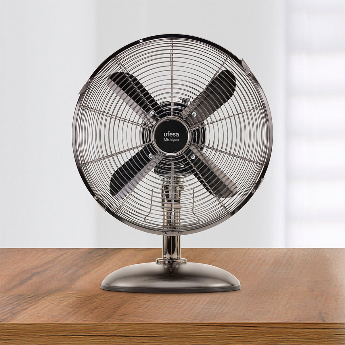 Un ventilador metálico Ufesa Michigan sobre una mesa de madera. Tiene cuatro aspas negras dentro de una jaula protectora. El fondo es una pared blanca.