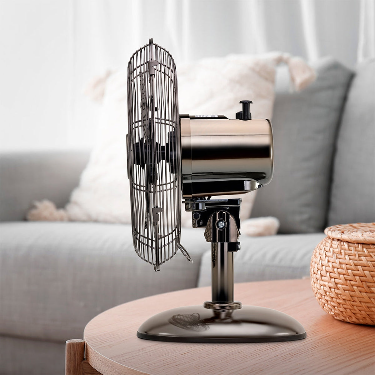 Un ventilador metálico sobre una mesa de madera. El fondo muestra un sofá y una cesta tejida. El ventilador está encendido.