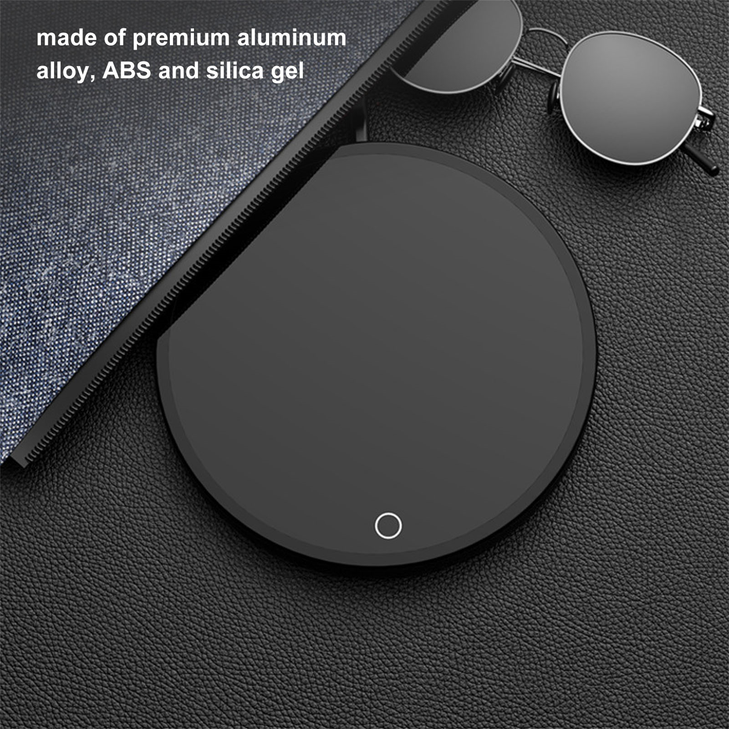 Schwarzes rundes Gerät mit einem kleinen weißen Kreis auf dunkler Oberfläche. Sonnenbrille und Tablet ebenfalls vorhanden.