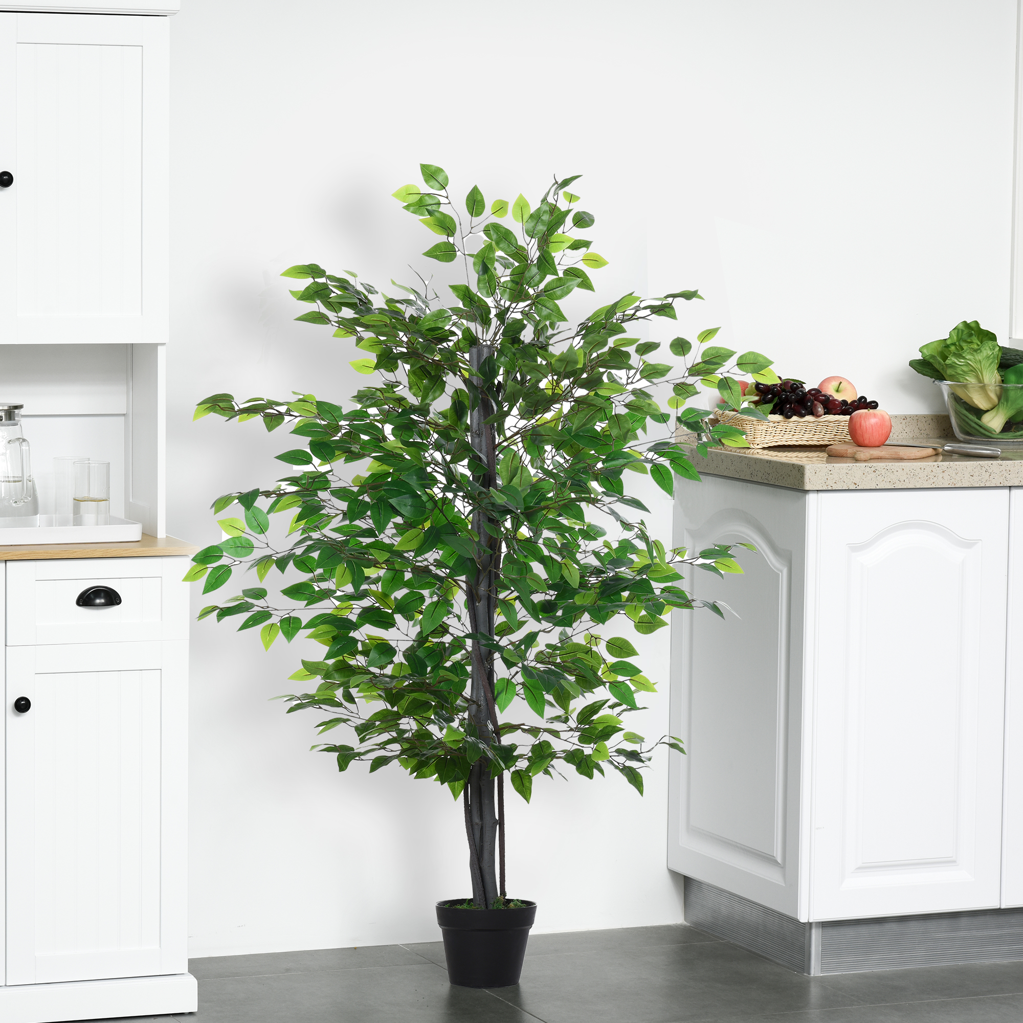 Ein hoher Kunstbaum in einem schwarzen Topf, vor weißen Küchenschränken platziert. Es gibt eine Arbeitsplatte mit Obst.