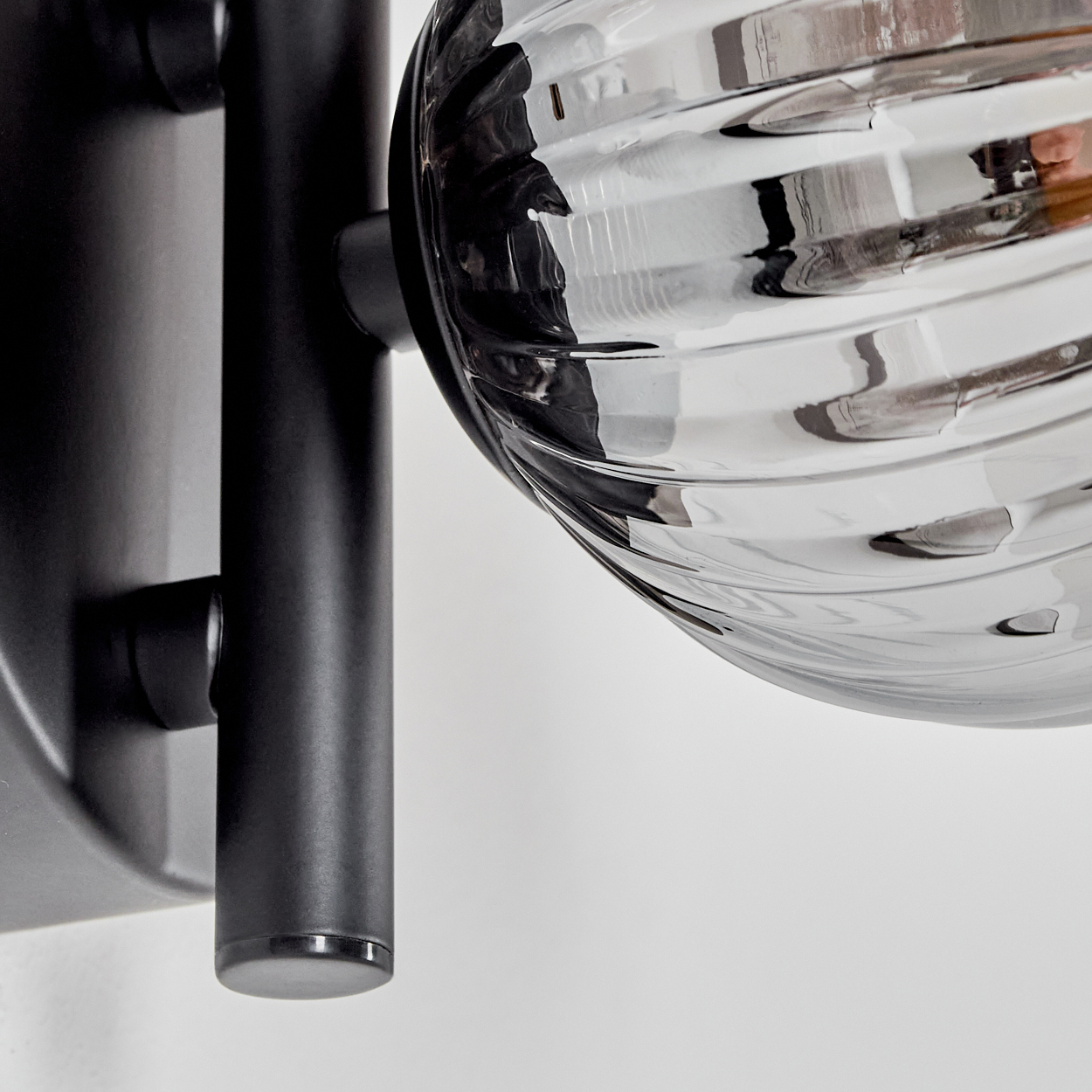 Nahaufnahme einer Wandlampe mit geripptem Glaskolben und schwarzem Metallarm. Die Lampe ist an einer weißen Wand befestigt.
