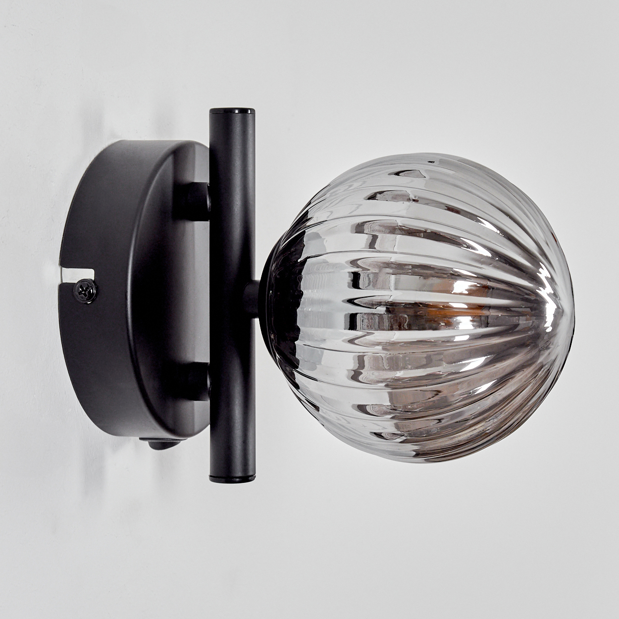 Eine Wandleuchte mit geripptem Glaskolben und schwarzem Metallarm. Die Lampe ist beleuchtet und an einer weißen Wand befestigt.
