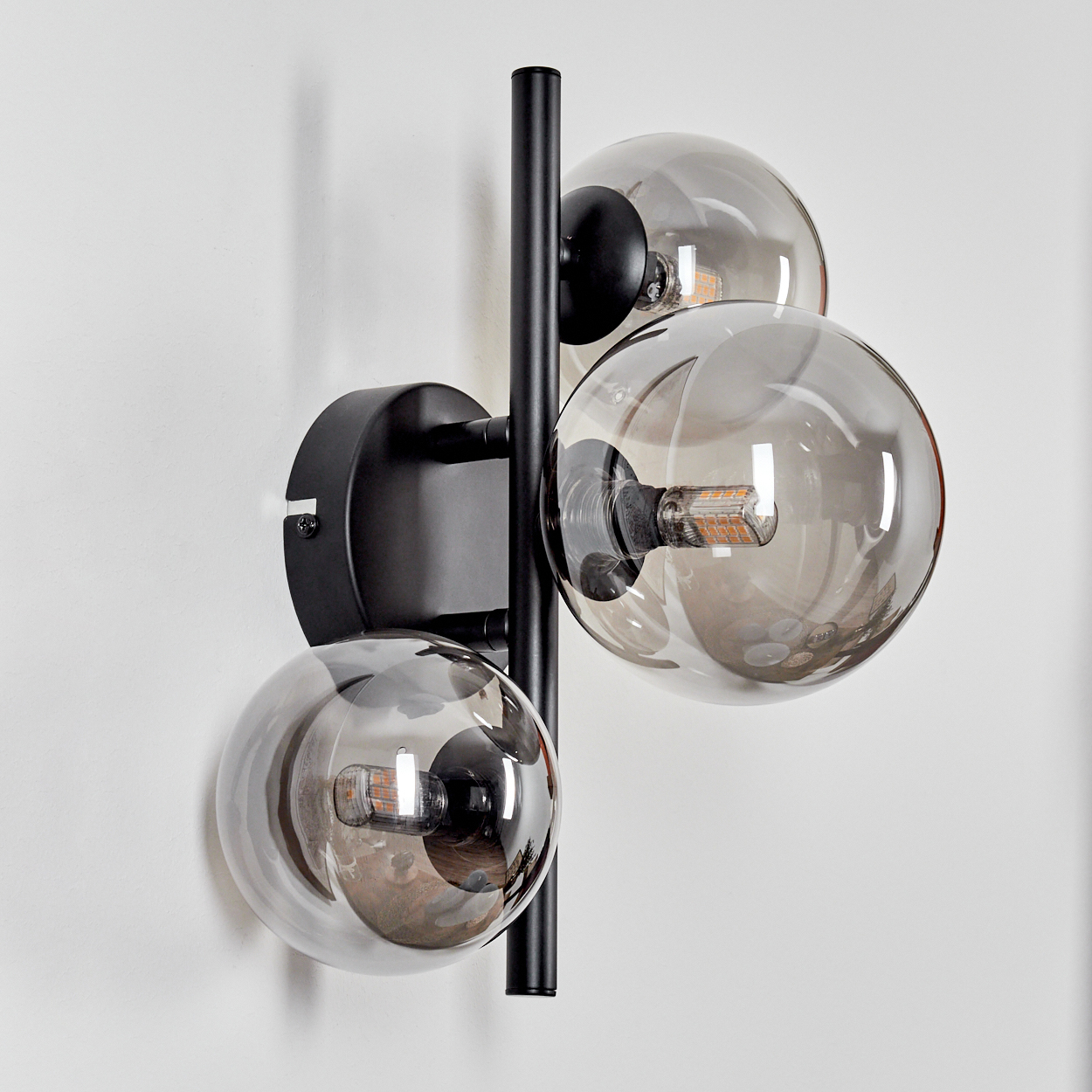 Eine schwarze Wandlampe mit drei rauchfarbenen Glaskugeln an einer weißen Wand.