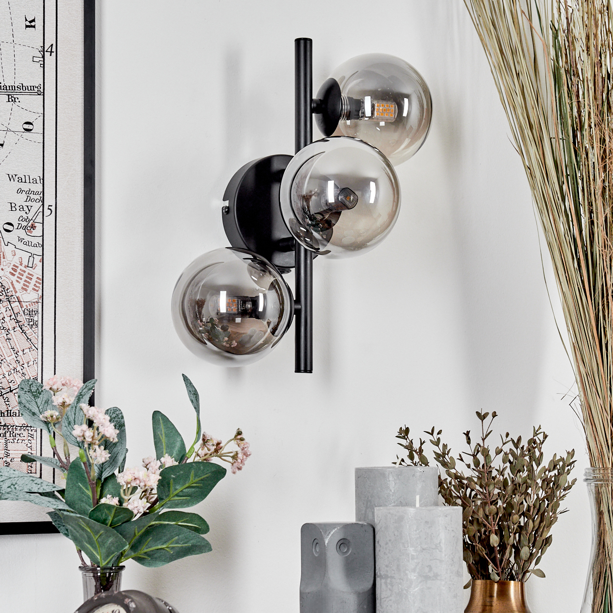 Wandlampe mit drei kugelförmigen Glasschirmen, schwarze Halterung. Blumen und Karte im Hintergrund.