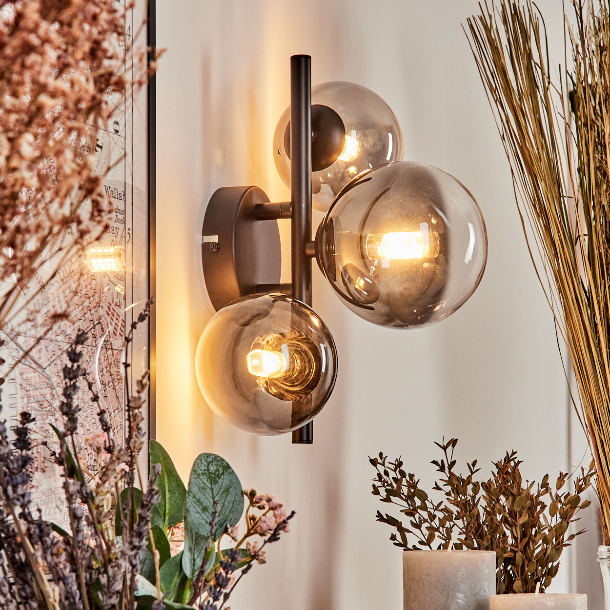 Wandlampe mit drei klaren Glaskugeln und schwarzem Rahmen, in der Nähe von Pflanzen, Karte und Kerzen.