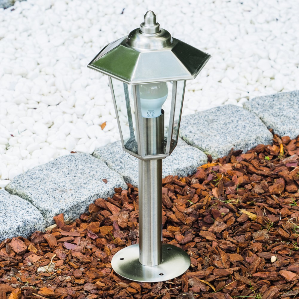 Eine silberne Gartenleuchte mit Glasoberteil, die in Rinde und weißem Stein steht.