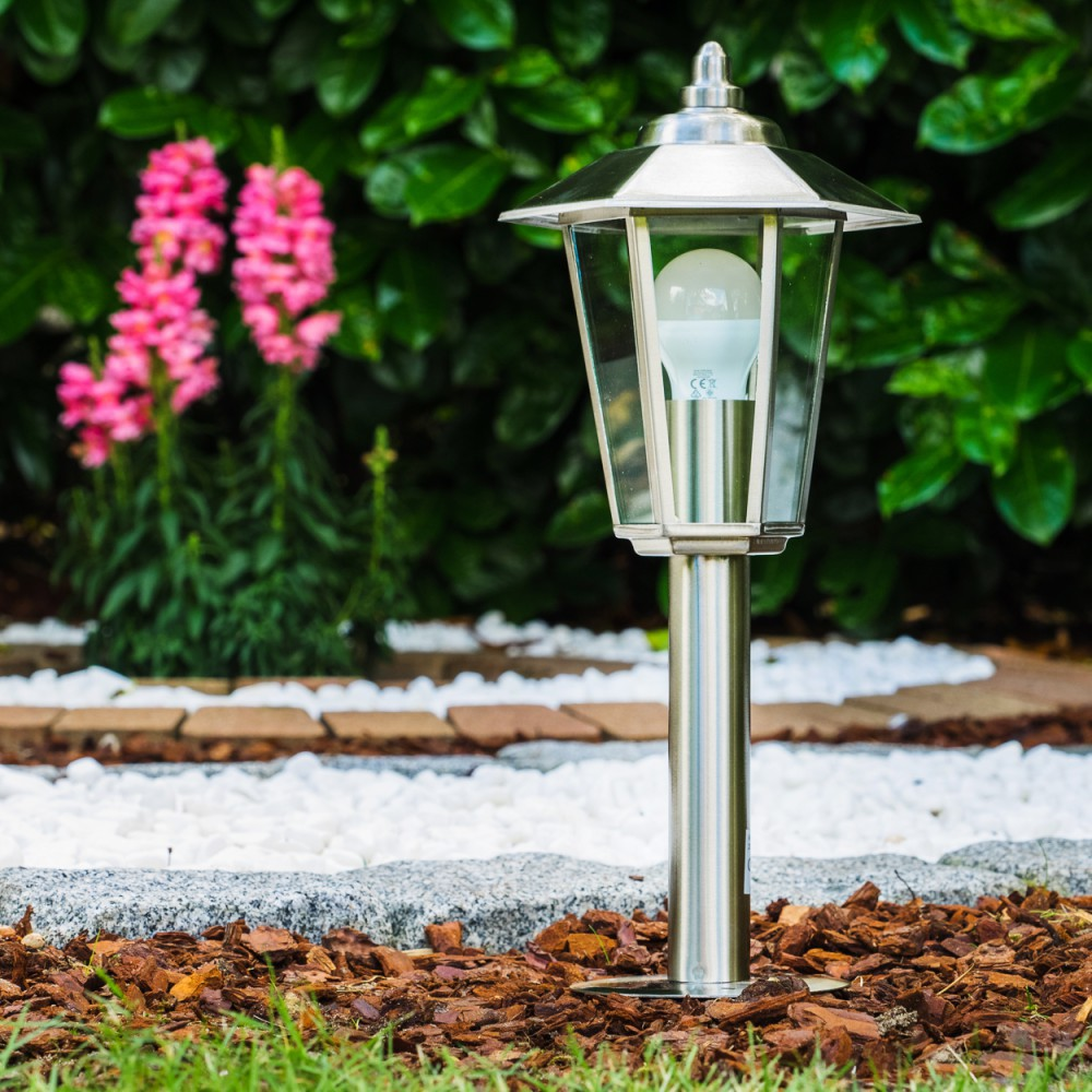 Eine silberne Lampenpfosten in einem Garten. Eine durchsichtige Lampe ist beleuchtet. Rosa Blumen und grüne Büsche sind sichtbar.